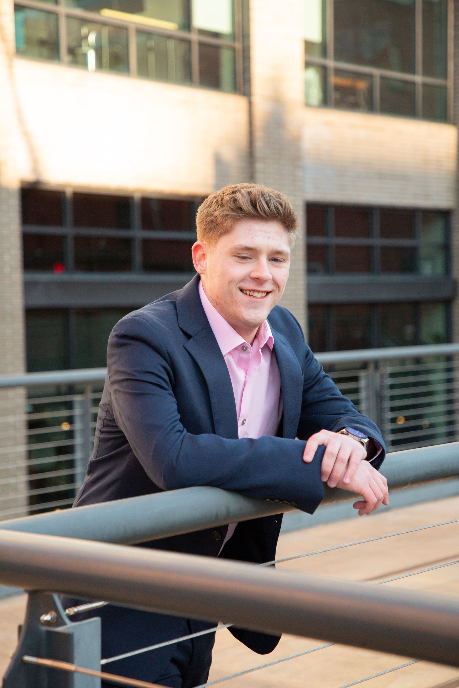 Smiling young man in navy suit and pink shirt leaning on a railing outside a modern building.
