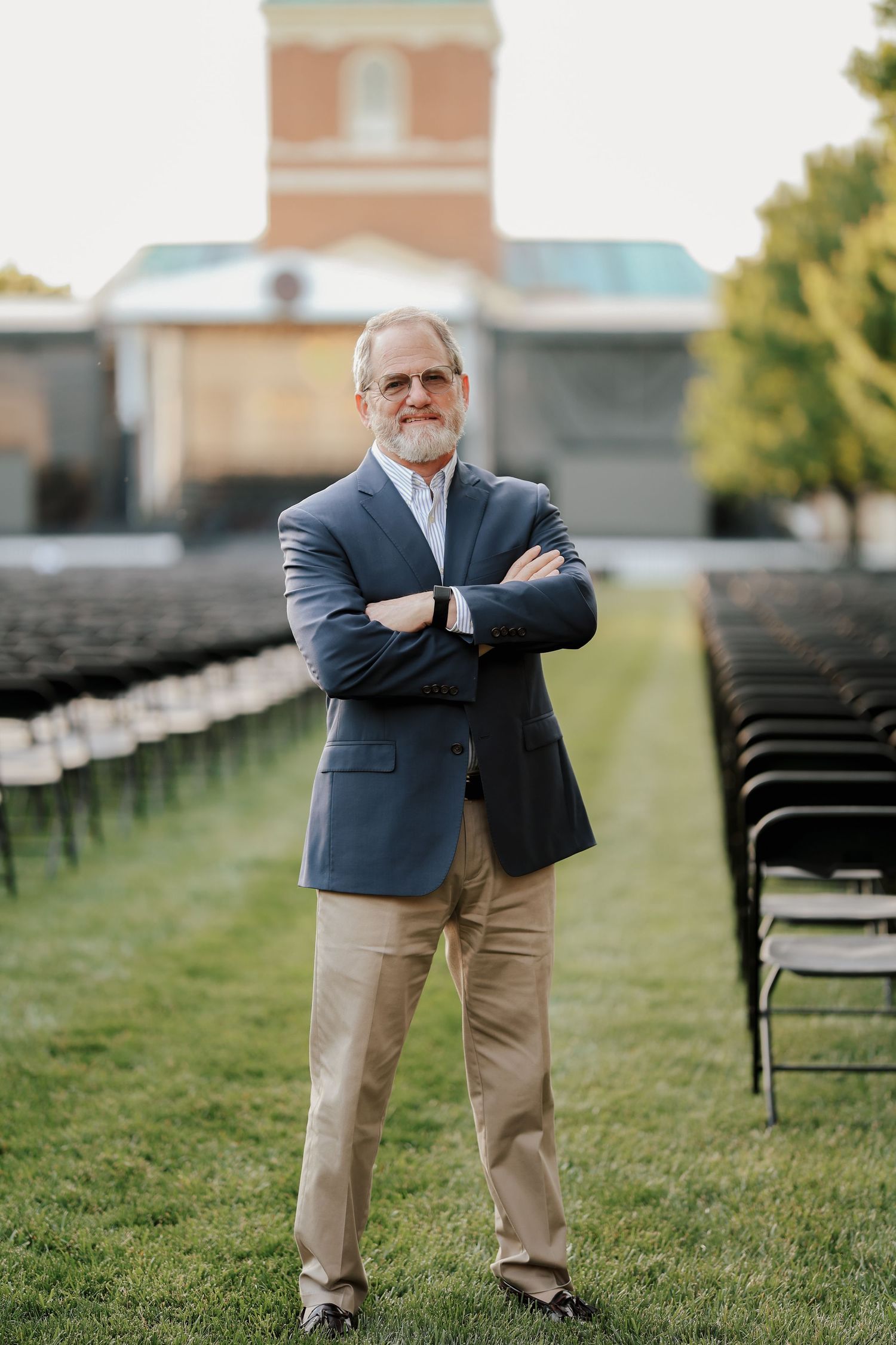 Distinguished older man in blue blazer standing with arms crossed on green lawn near brick building.