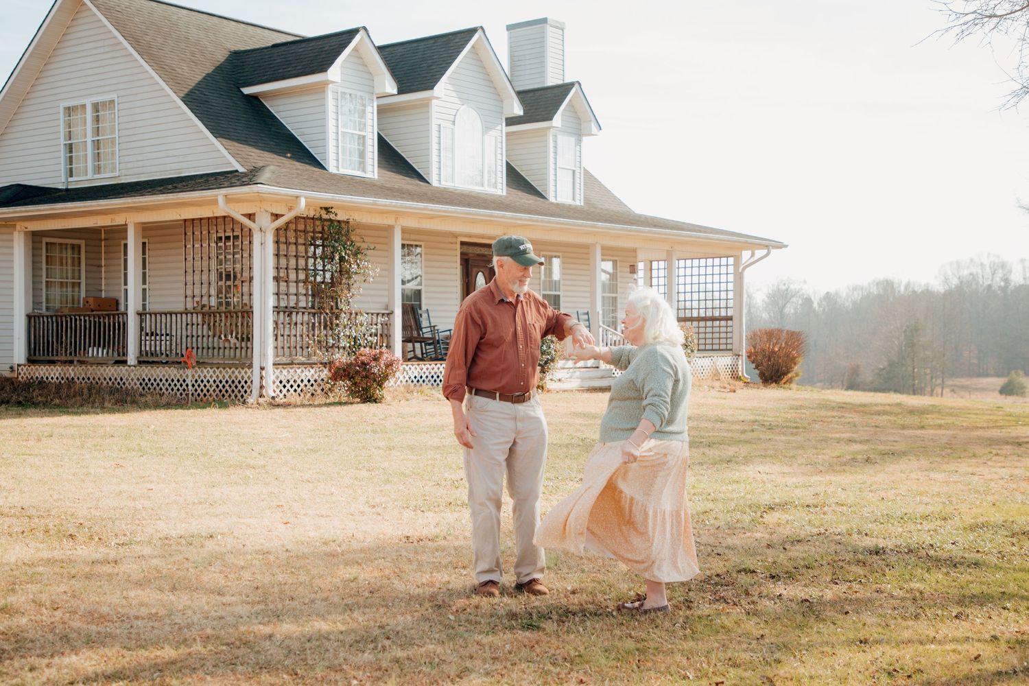 Senior couple holding hands and dancing playfully in front of a white country farmhouse.