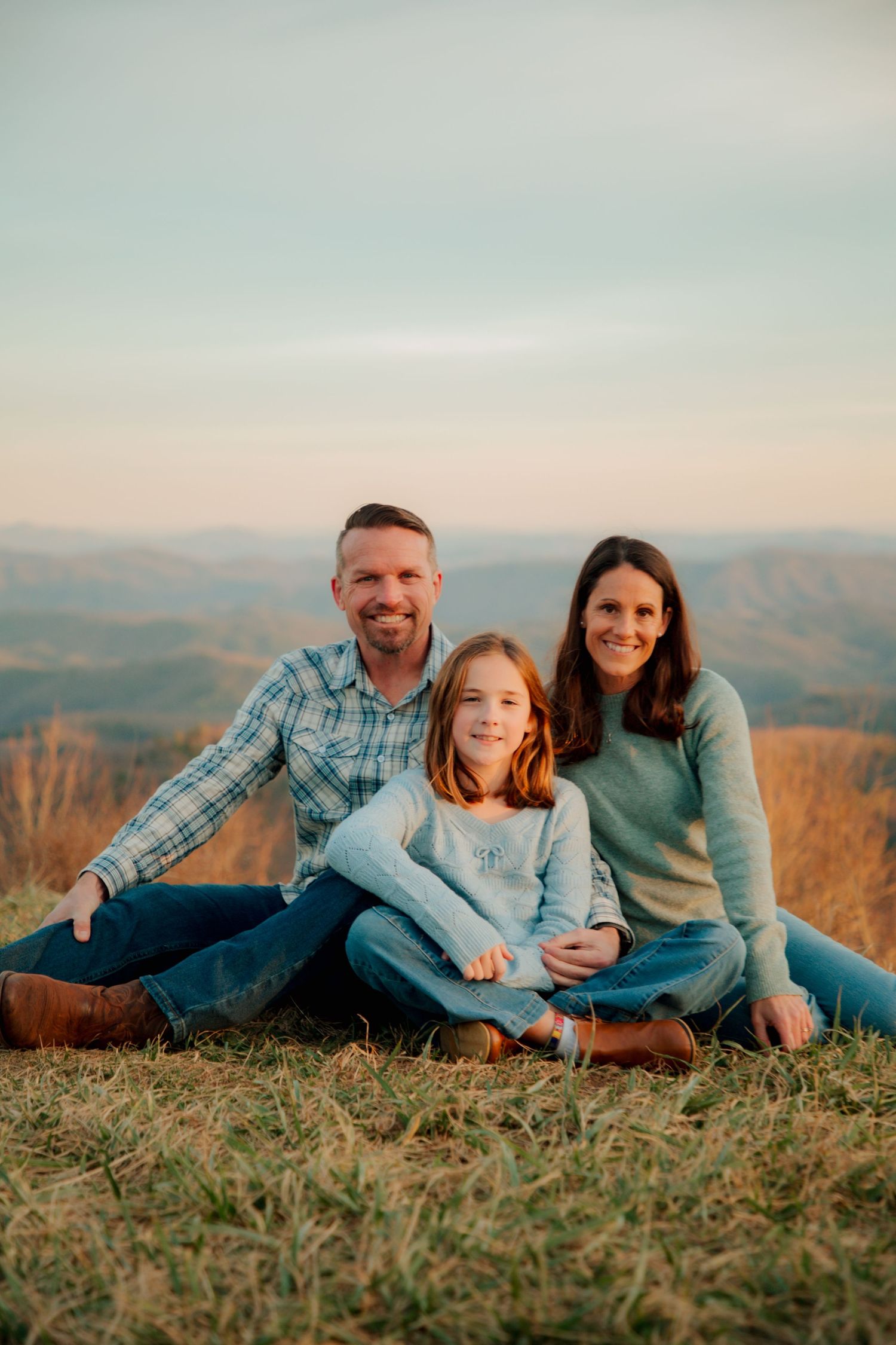 A smiling family of three poses together on a hilltop with sweeping mountain views under a soft evening sky.