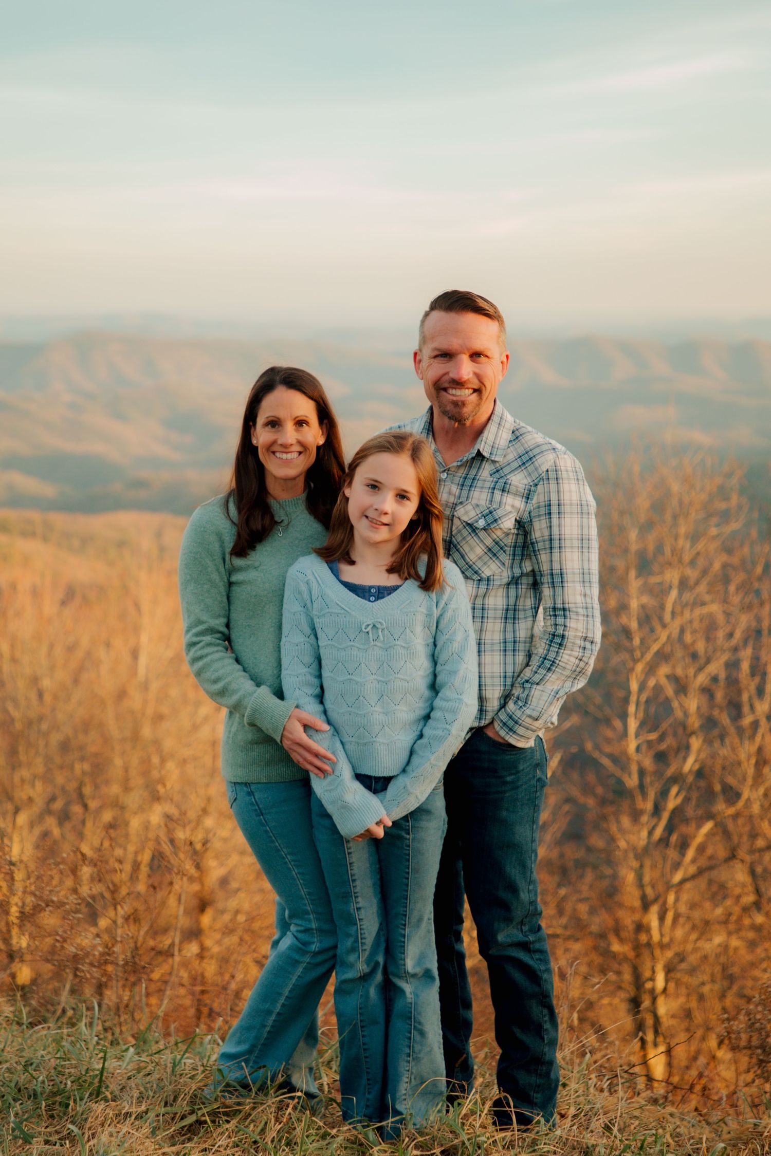Family of three stands smiling together with colorful autumn mountain scenery behind them.