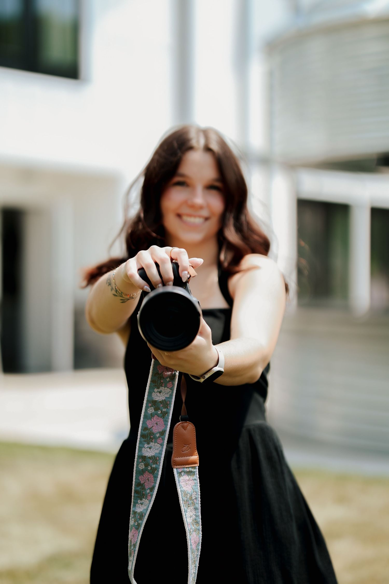 Smiling photographer in black dress extends camera with floral strap toward viewer outdoors.