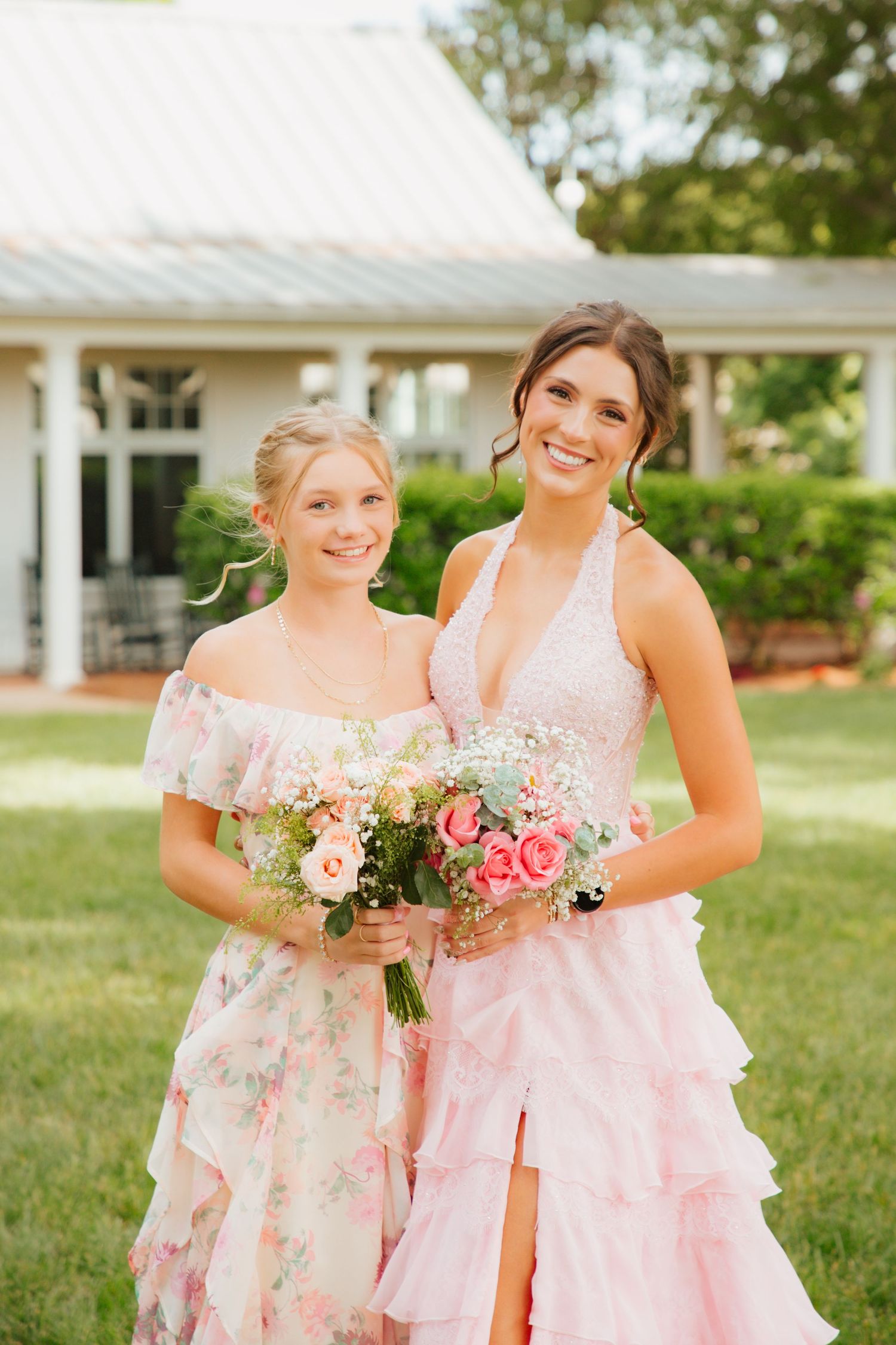 Two smiling young women in pink floral and pink ruffle formal gowns holding bouquet on green lawn.
