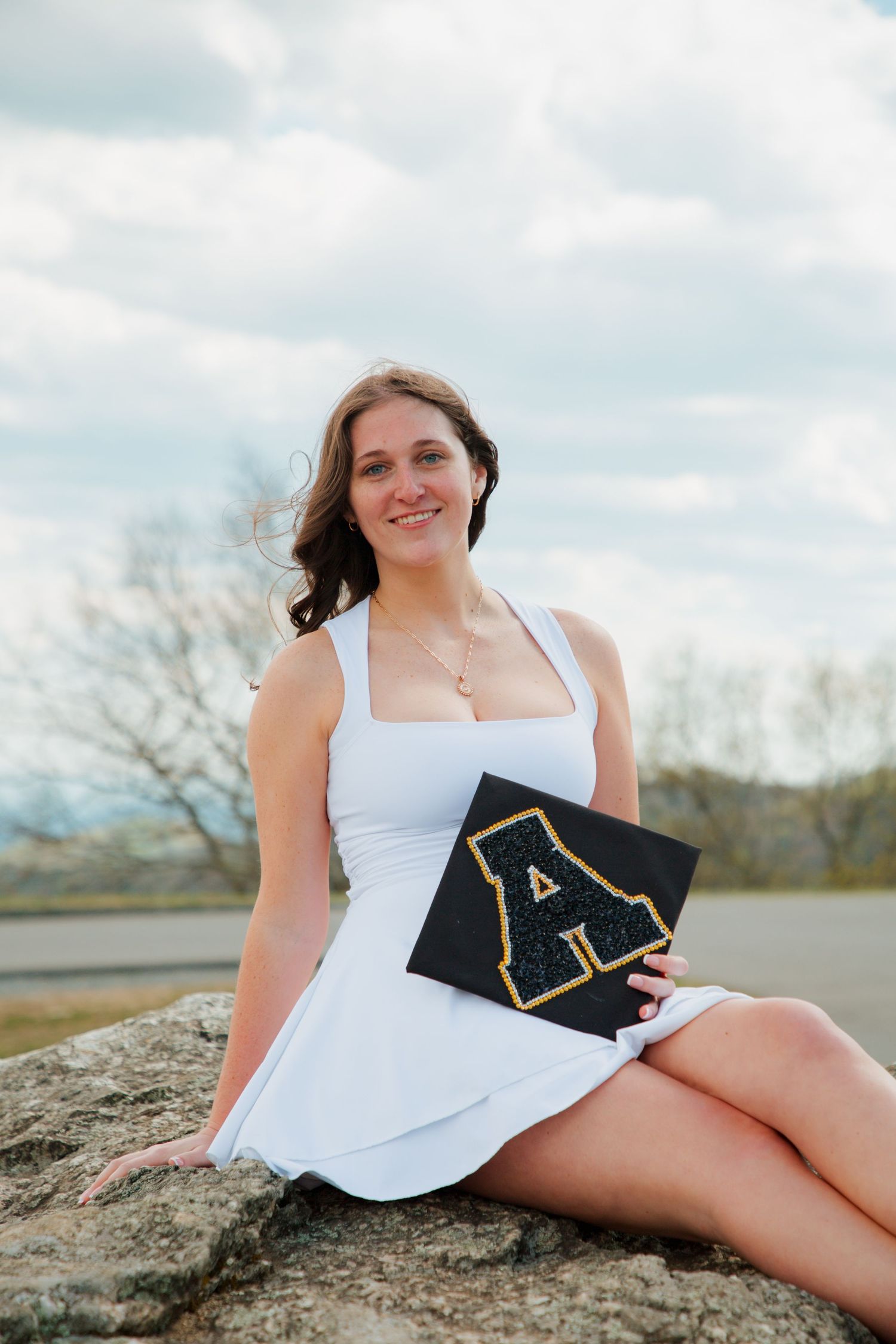 Smiling young woman in white dress holding Appalachian State graduation cap while seated on rocks outdoors.