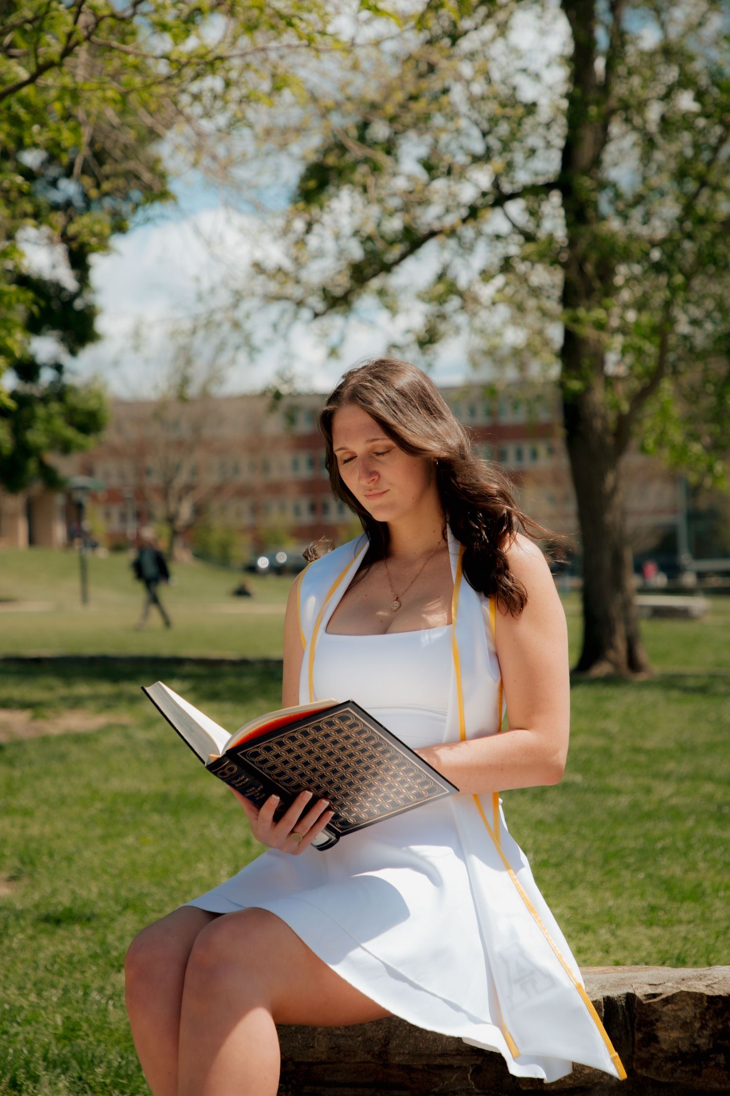 Female graduate in white dress sitting on log reading diploma in sunny campus park setting.