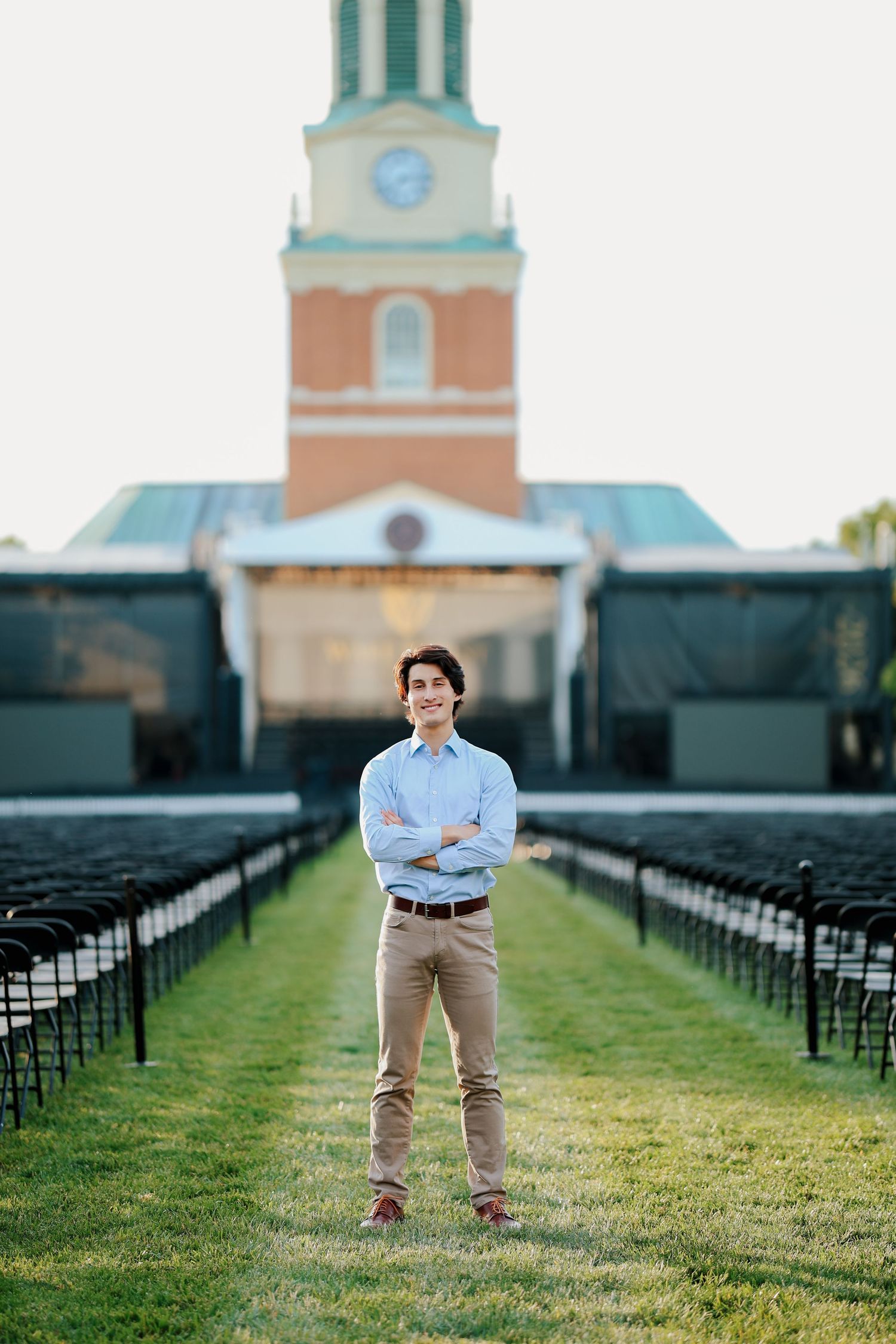 Smiling young man in blue shirt stands with crossed arms on lawn between rows of chairs.