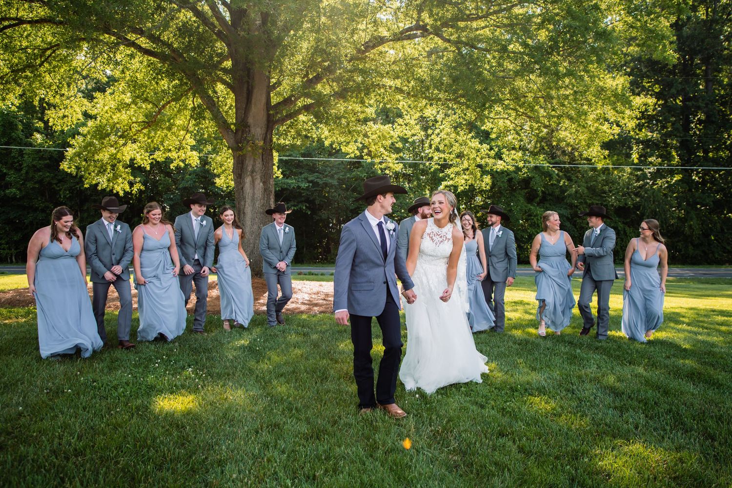 Bride and groom pose together on sunlit lawn as wedding party stands casually behind them near large tree.