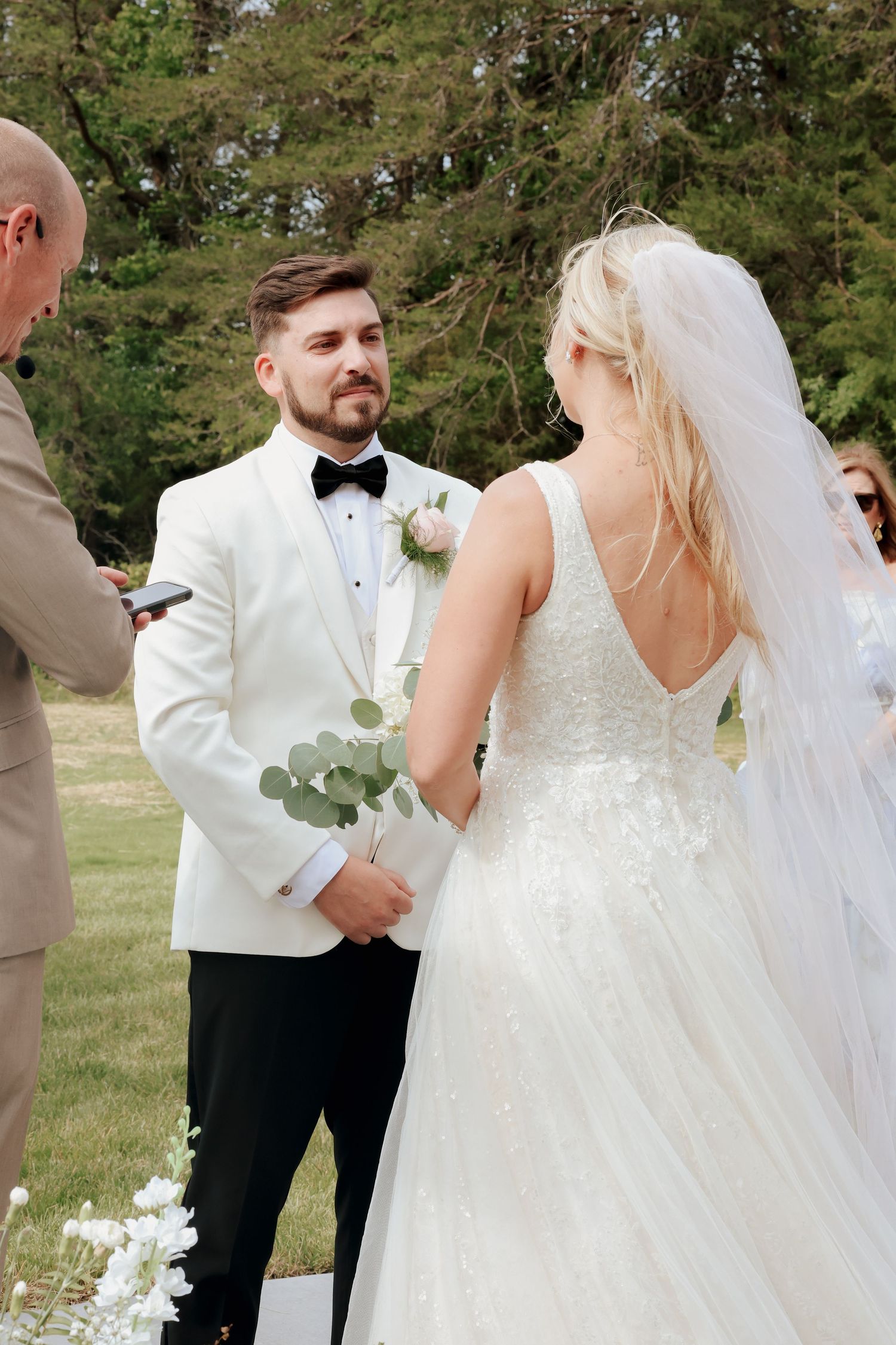 Groom in white tuxedo gazes lovingly at bride during outdoor wedding ceremony with wooden cross.