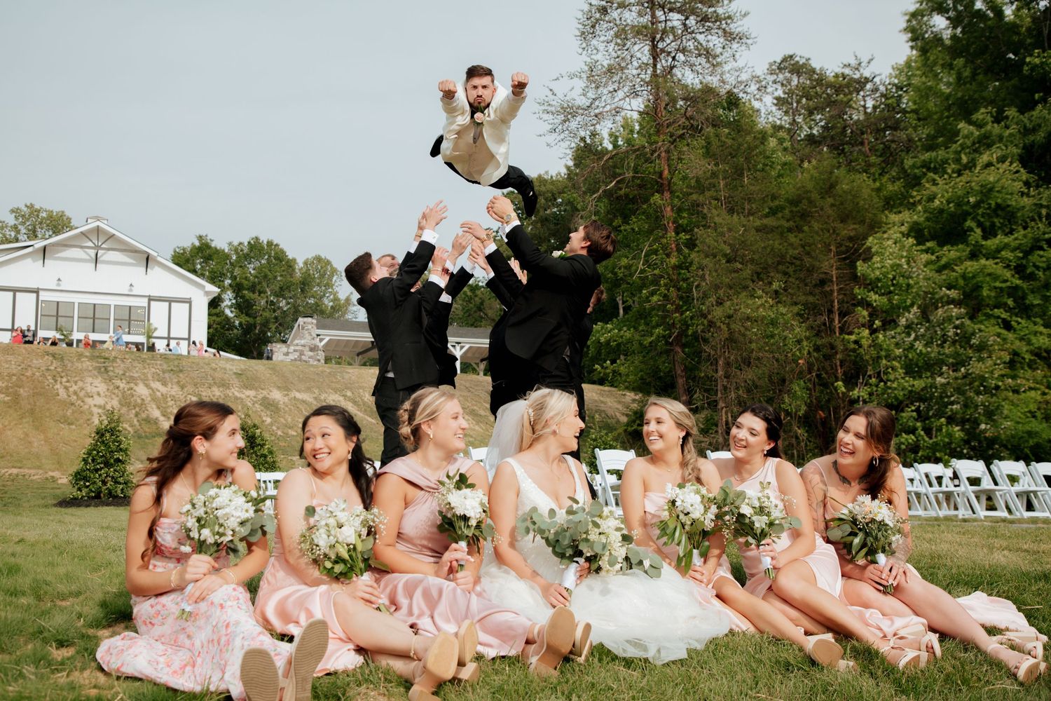 Groomsmen tossing groom in air while bridesmaids in blush dresses laugh on grass.