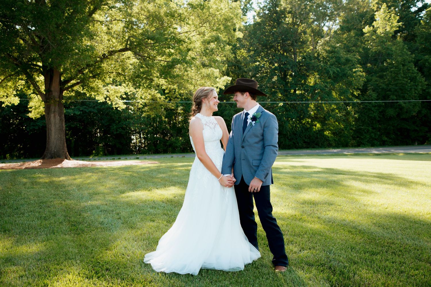 Bride in white lace gown and groom in gray suit holding hands on green lawn.