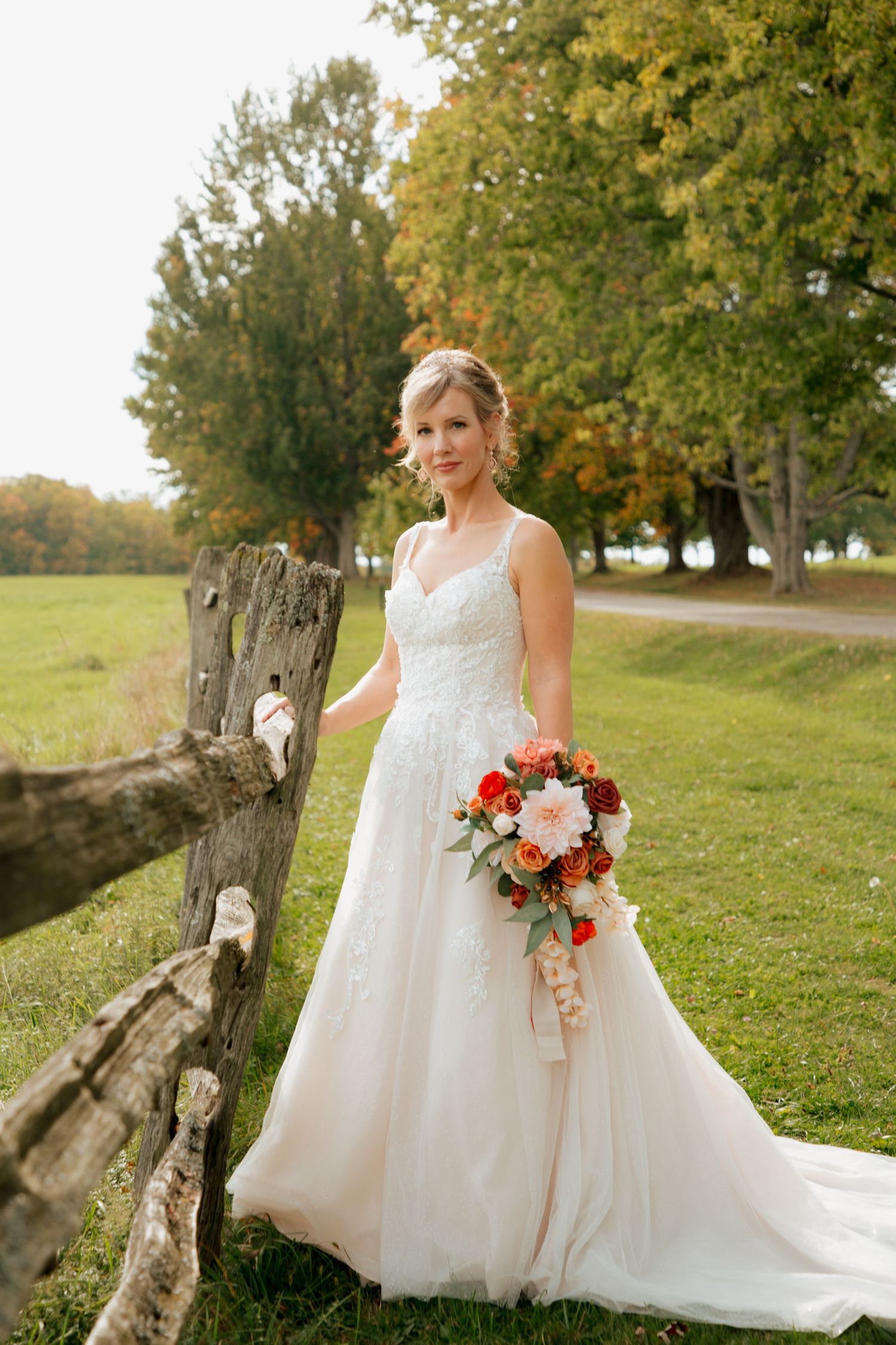 Bride in lace wedding gown holding orange and white bouquet posing beside a weathered wooden fence.