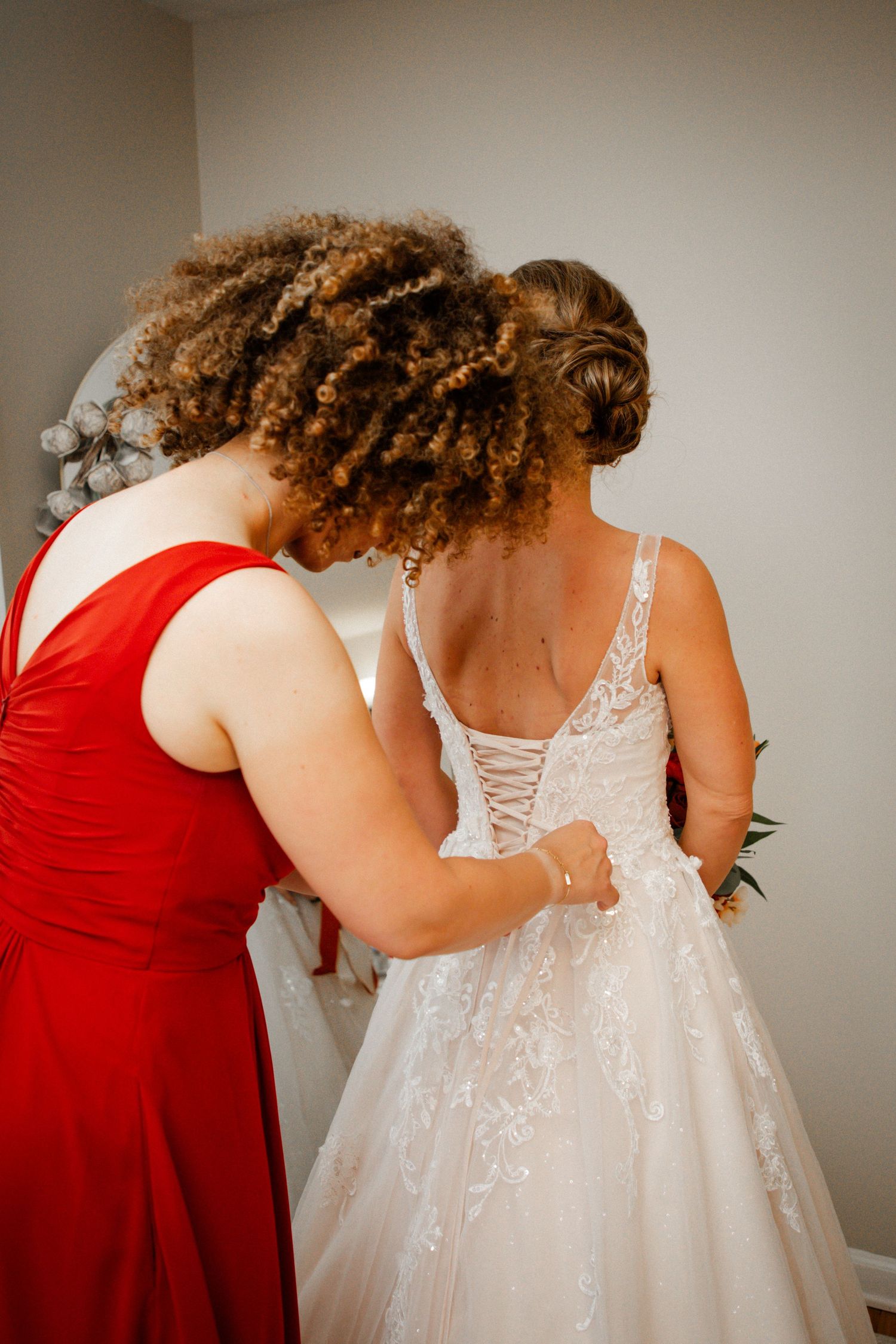 Bridesmaid in red dress lacing up bride's corset back wedding gown.
