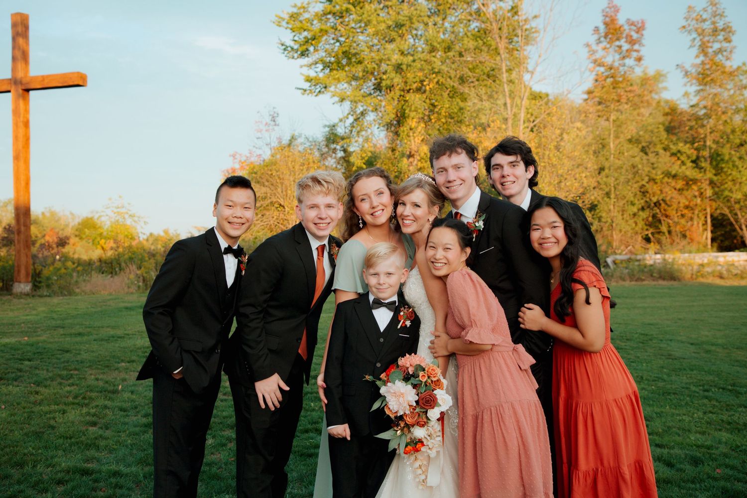 Bride and groom with a group of young guests smiling near a wooden cross in autumn.