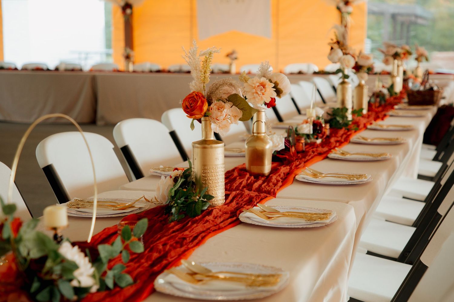 Long wedding reception table with red velvet runner, gold vases with floral centerpieces, and white folding chairs.