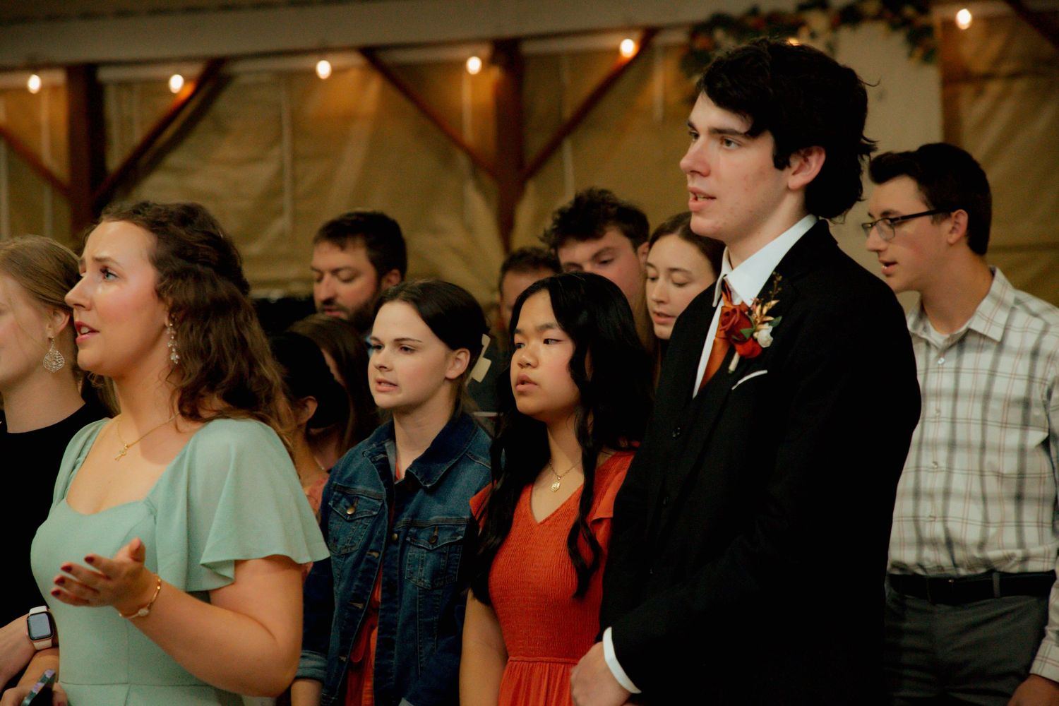 Young wedding guests in formal attire stand together during a ceremony in a warmly lit barn.