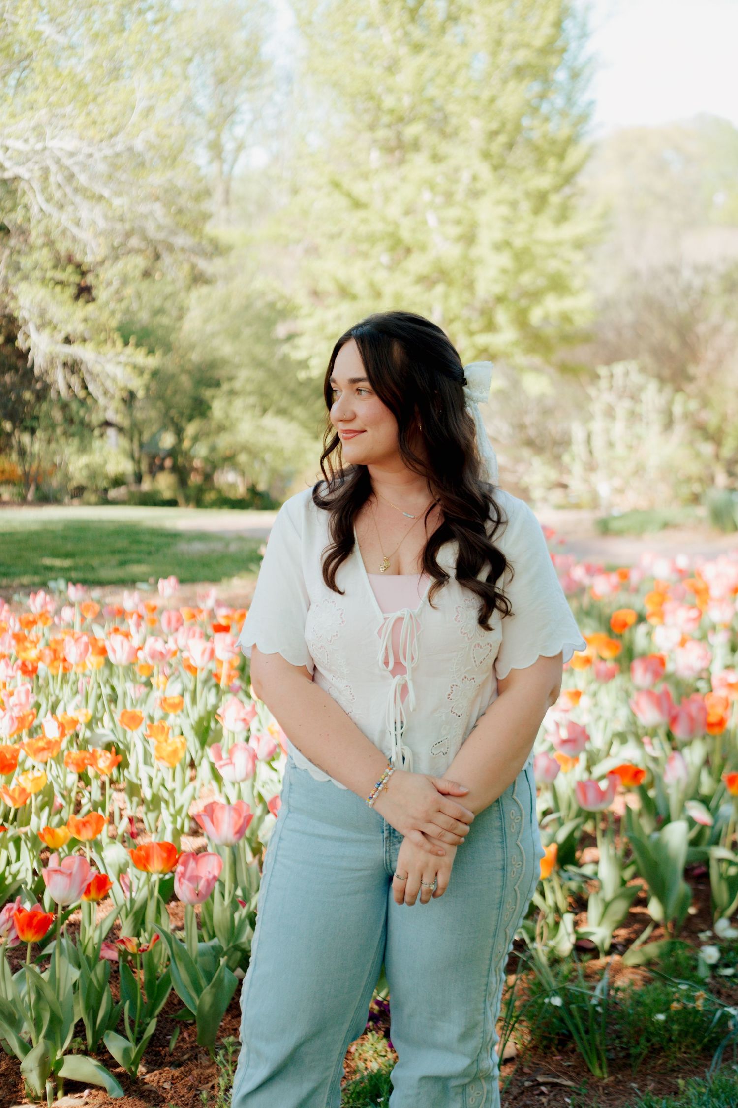 Young woman in white top and light blue jeans stands among colorful tulips in sunny garden.