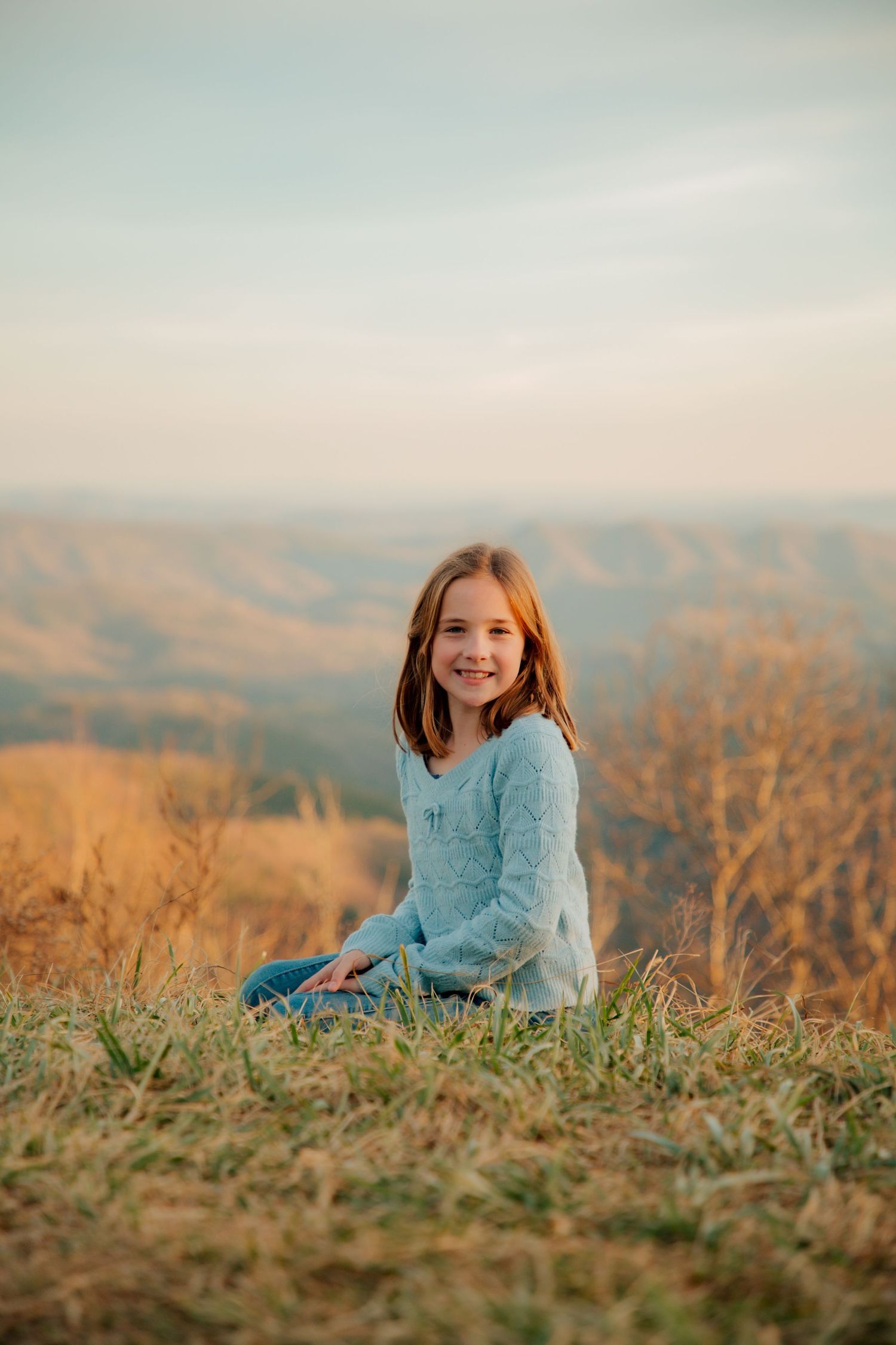 Young girl in blue sweater sits in grass smiling with scenic mountain hills behind her.