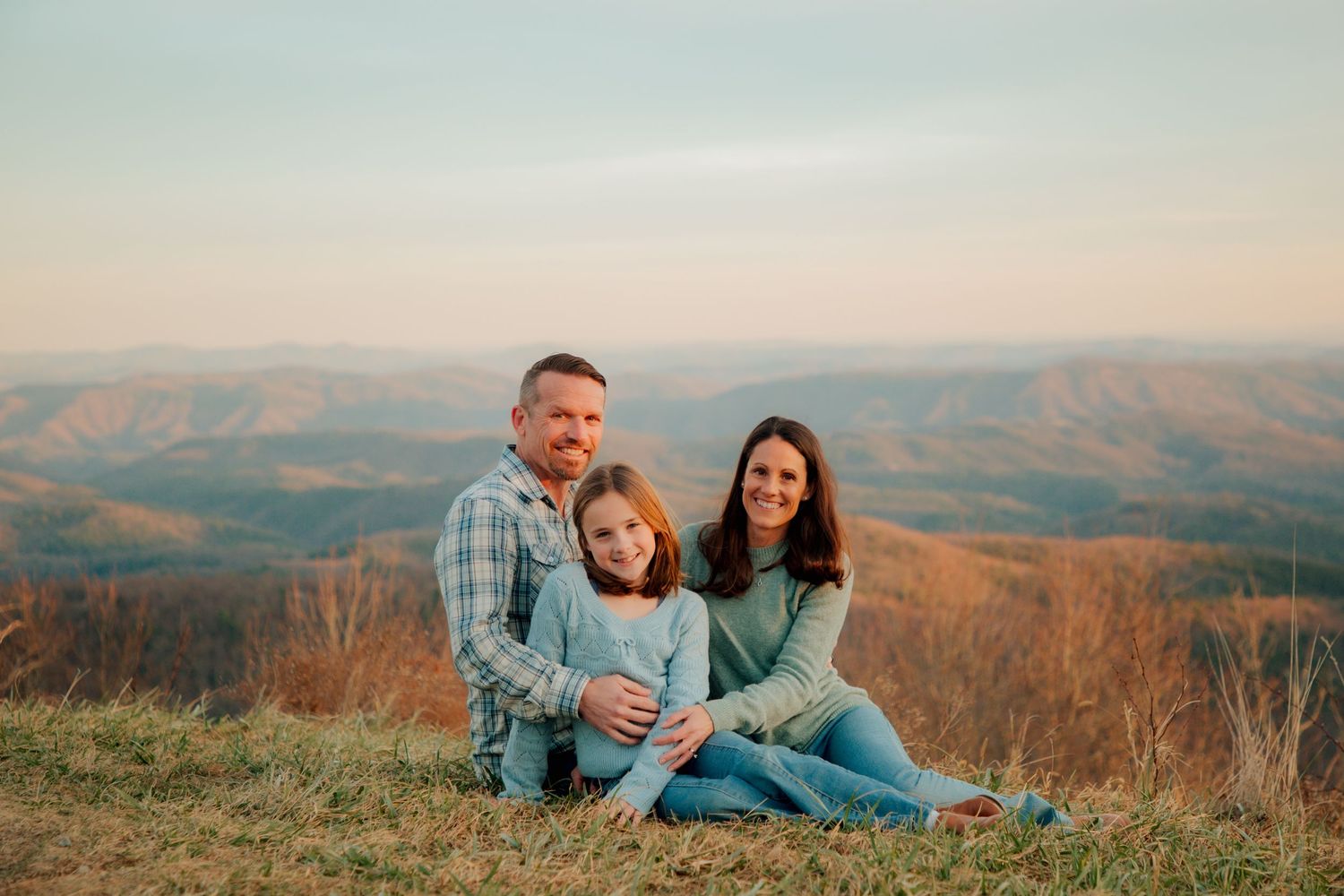 A family of three poses warmly together on a scenic mountain overlook during a beautiful golden hour sunset.