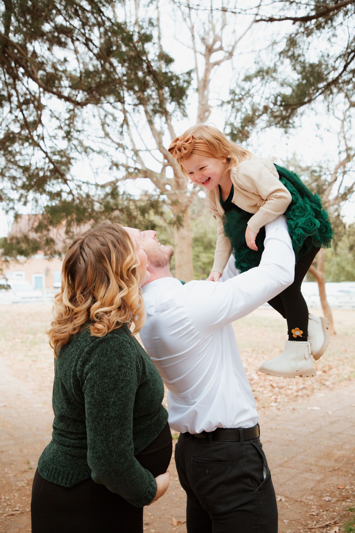 Parents playing with laughing toddler daughter in green dress during outdoor fall family session.