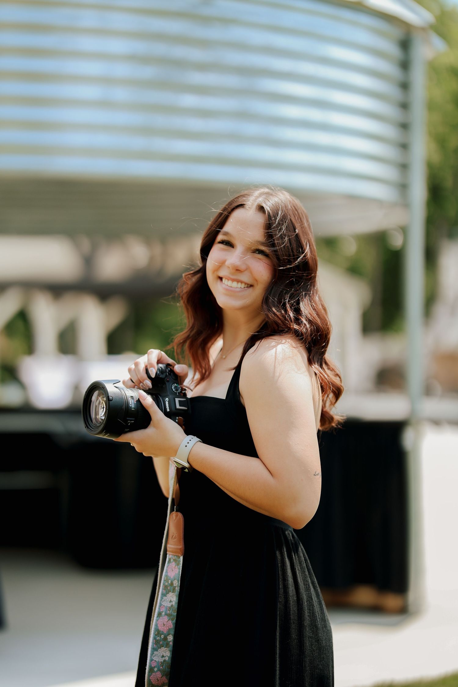 Smiling female photographer in black dress holds camera with floral strap outdoors in sunlight.