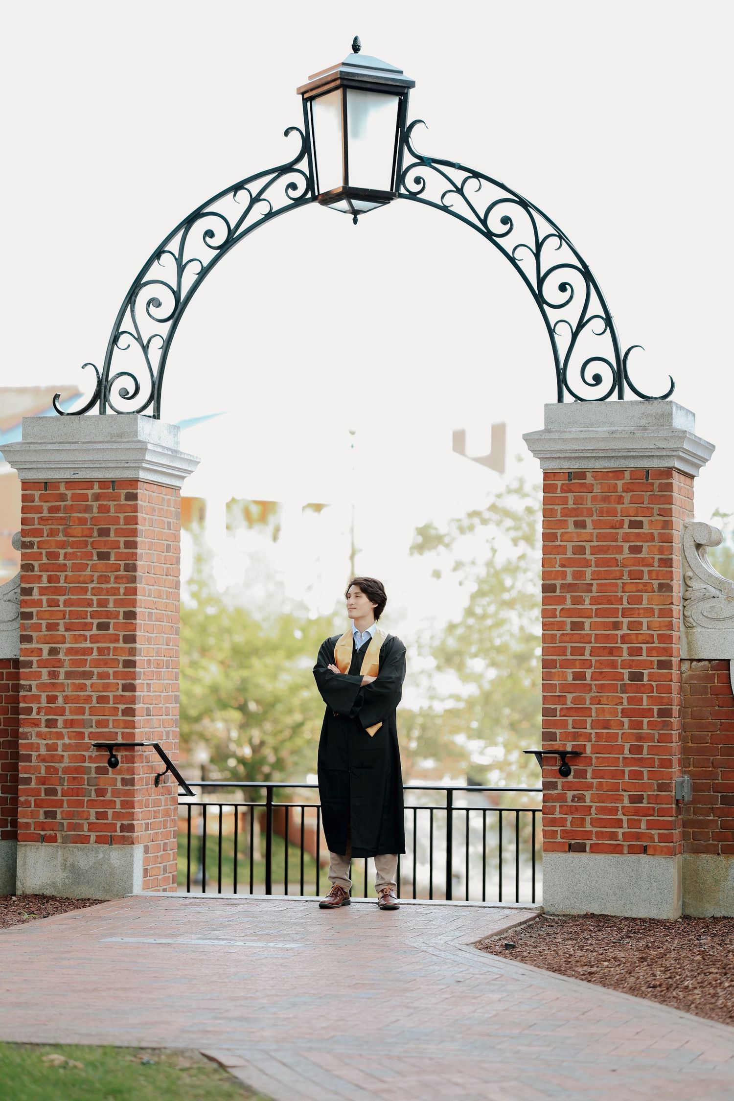 Graduate in black cap and gown stands under ornate iron arch at brick campus entrance.