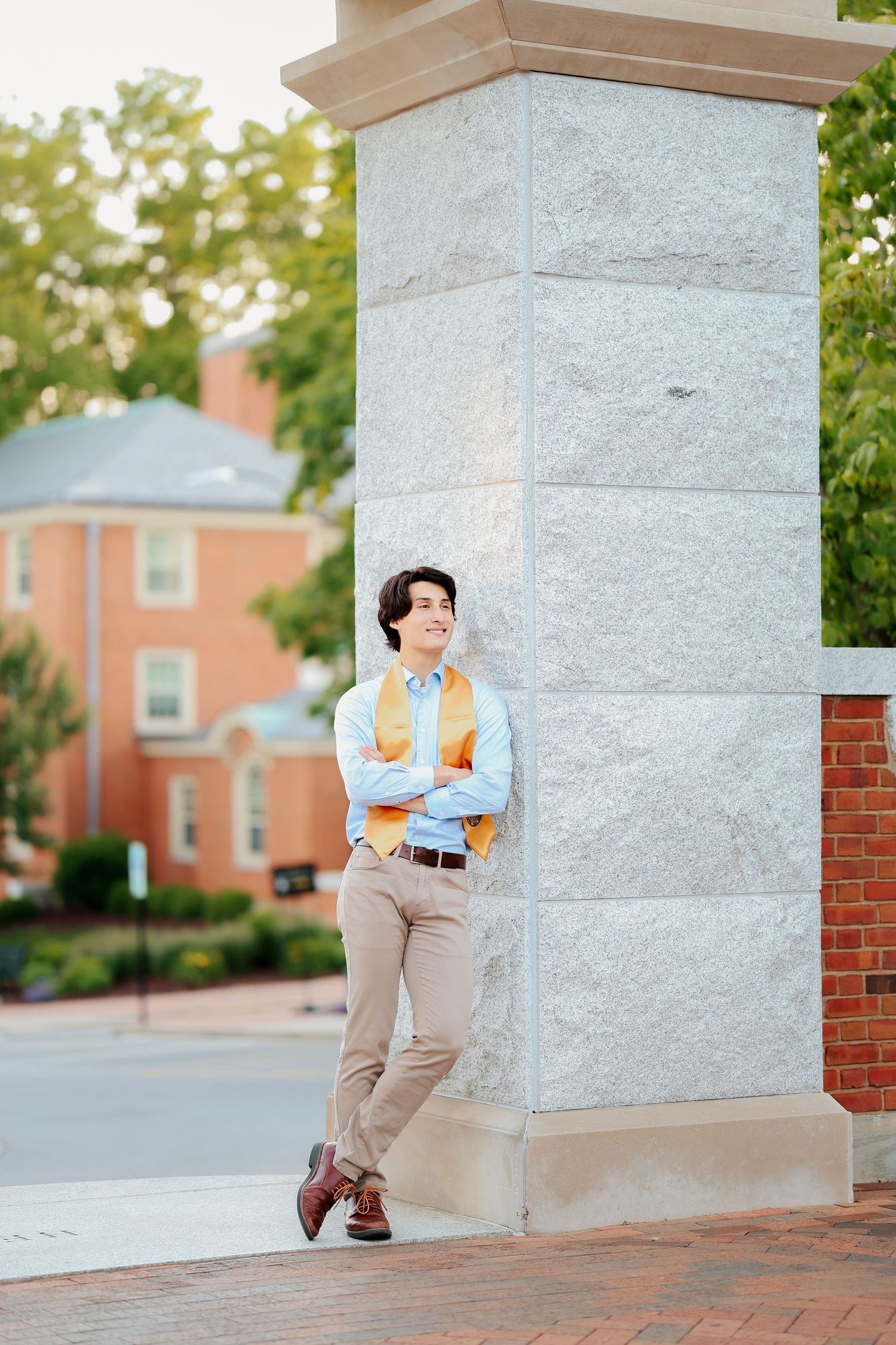 Smiling graduate in blue shirt and gold stole leans against stone pillar on brick campus.