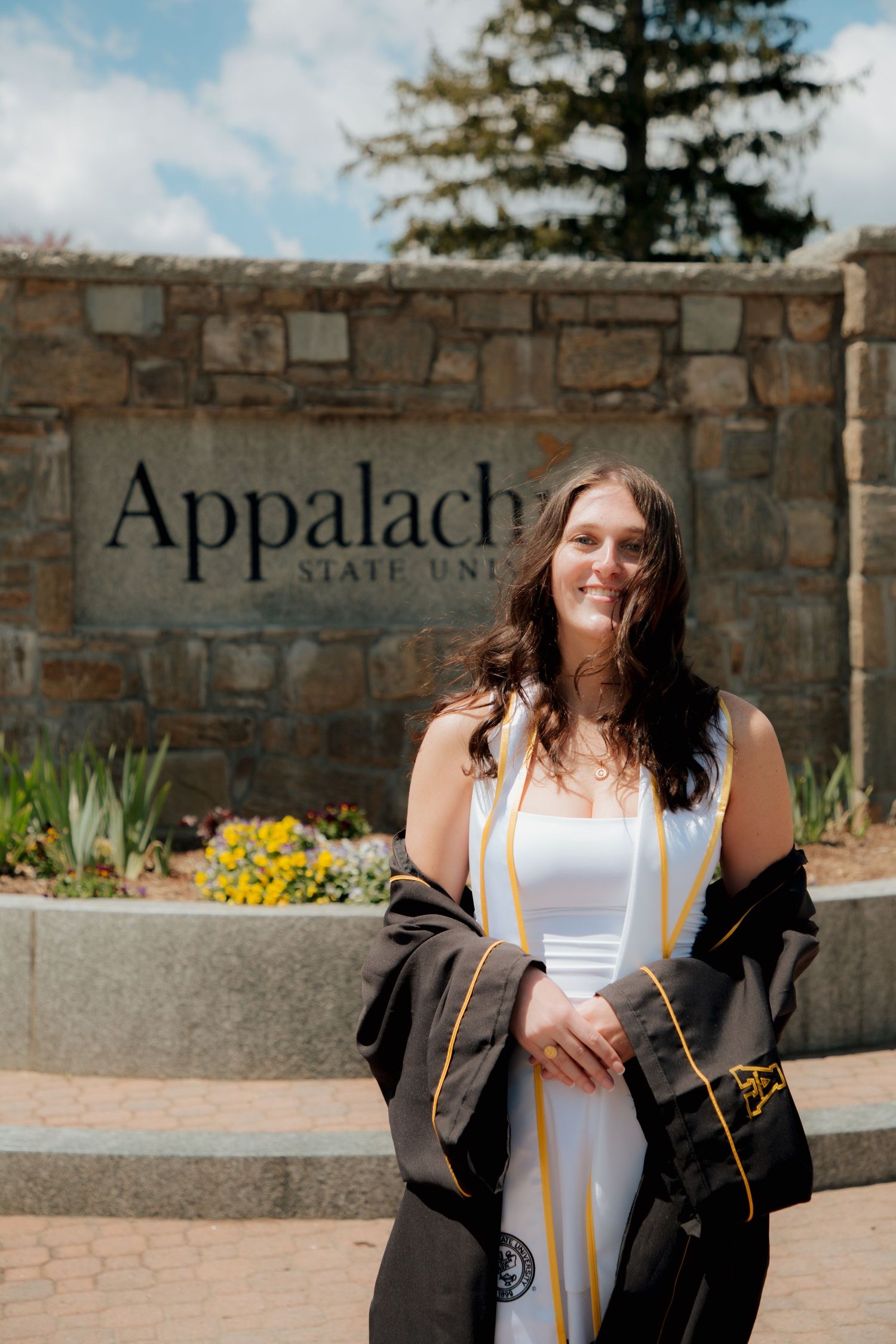 Smiling female graduate in black gown standing before Appalachian State University stone sign.