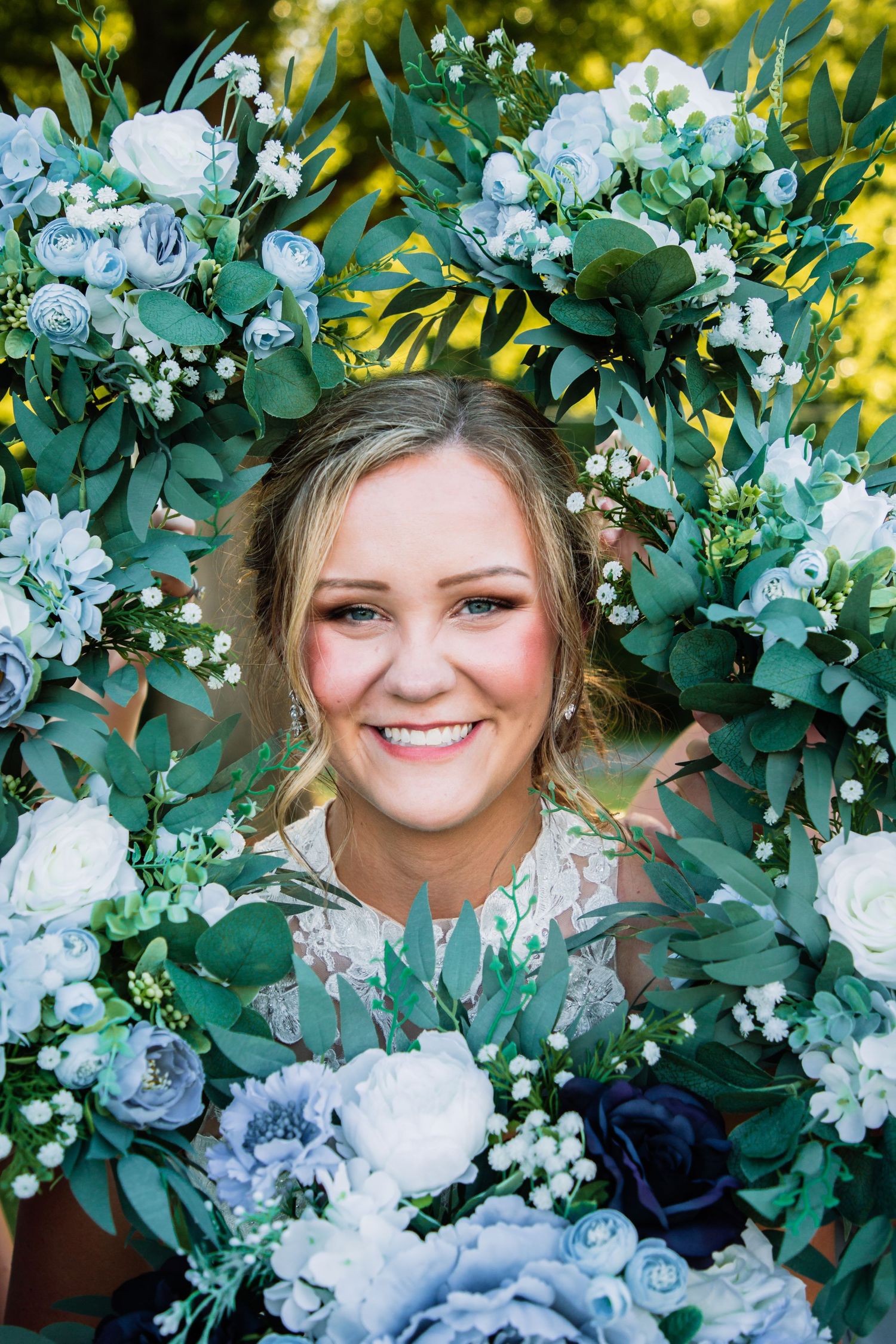 Smiling bride framed by lush blue and white floral arrangement with green eucalyptus leaves in a close-up portrait.