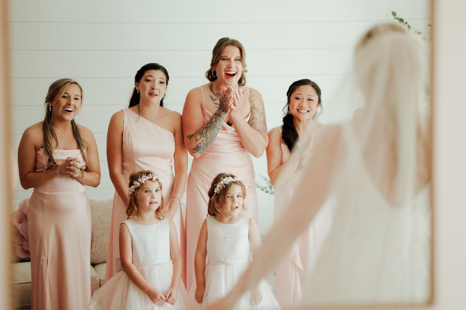 Bridesmaids in blush gowns and flower girls react with excitement to bride during first look.