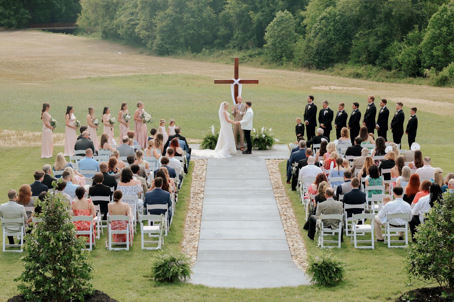 Aerial view of elegant outdoor wedding ceremony with wooden cross, bridesmaids, groomsmen, and seated guests.