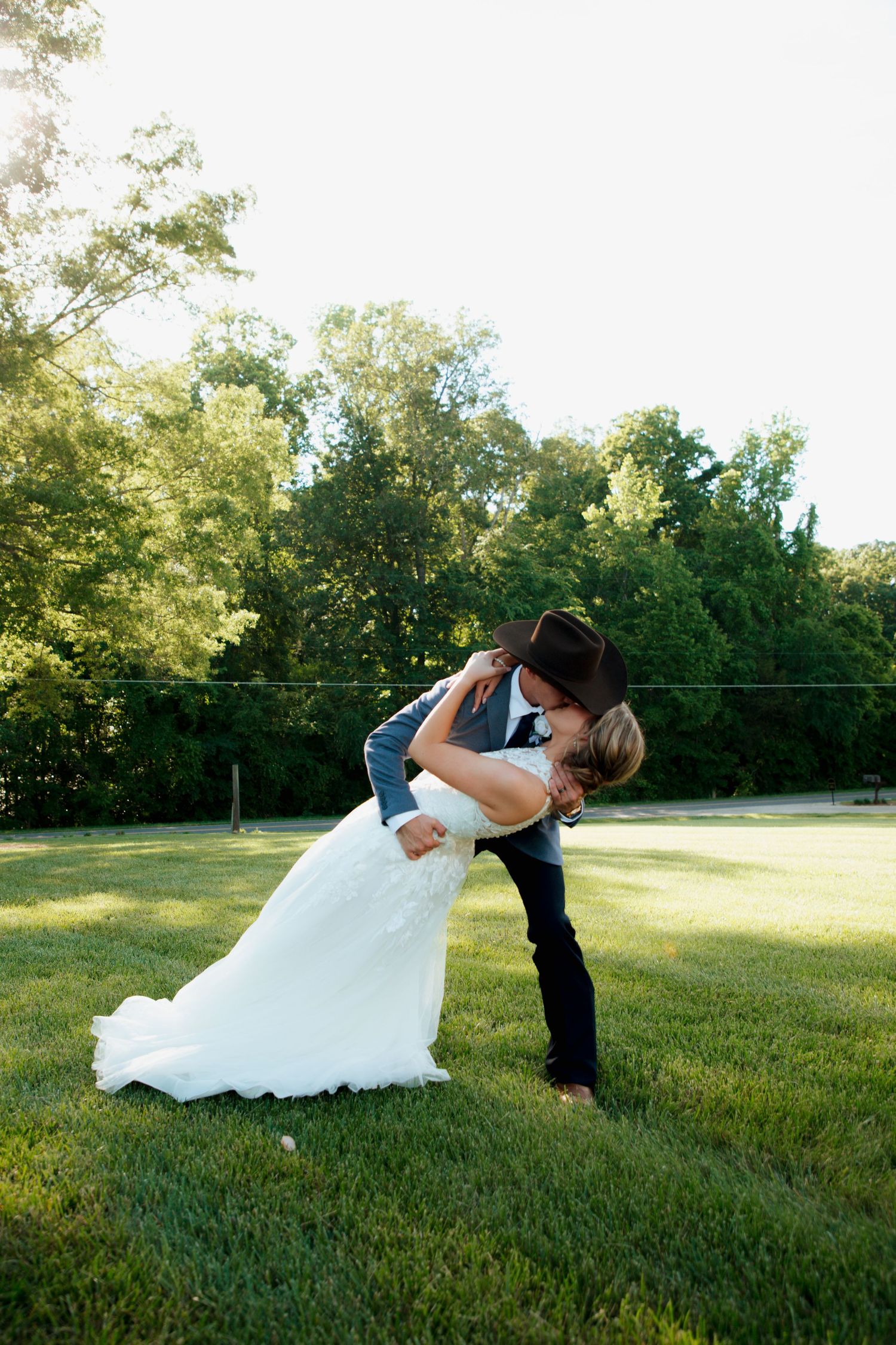 Groom in cowboy hat dipping bride in white gown for a kiss in golden sunlight outdoors.