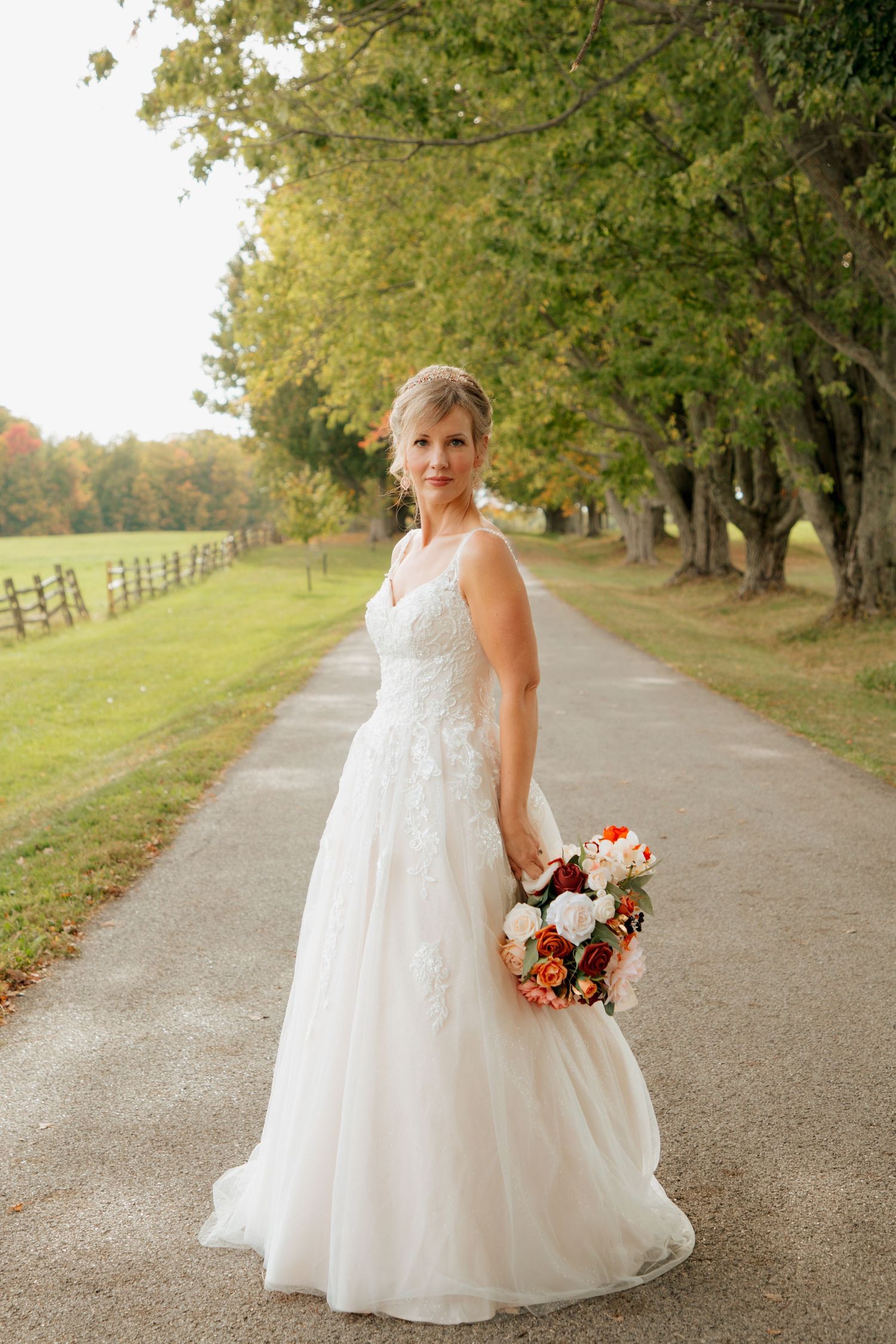 Bride in elegant white ball gown holding fall bouquet standing on a scenic tree-lined driveway.