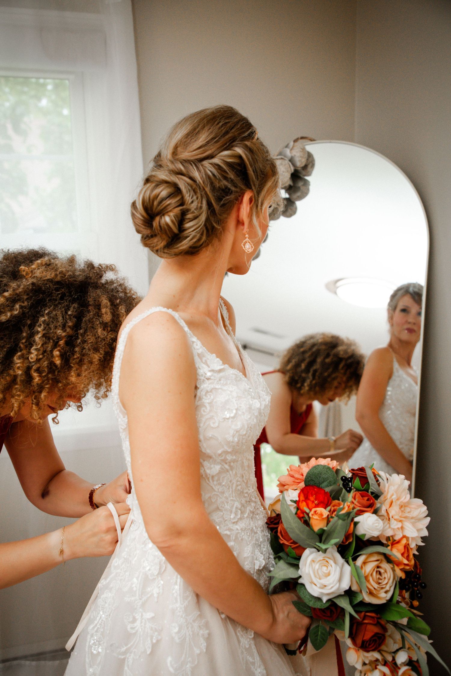 Bride holding autumn bouquet while bridesmaid fastens lace wedding gown before mirror.