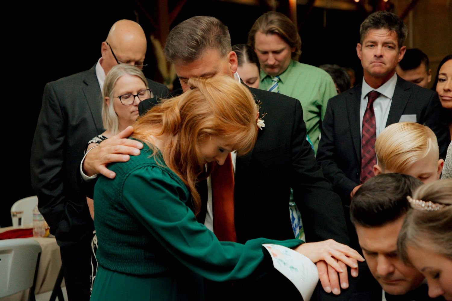 Wedding guests in formal attire gather closely during an emotional prayer moment at reception.