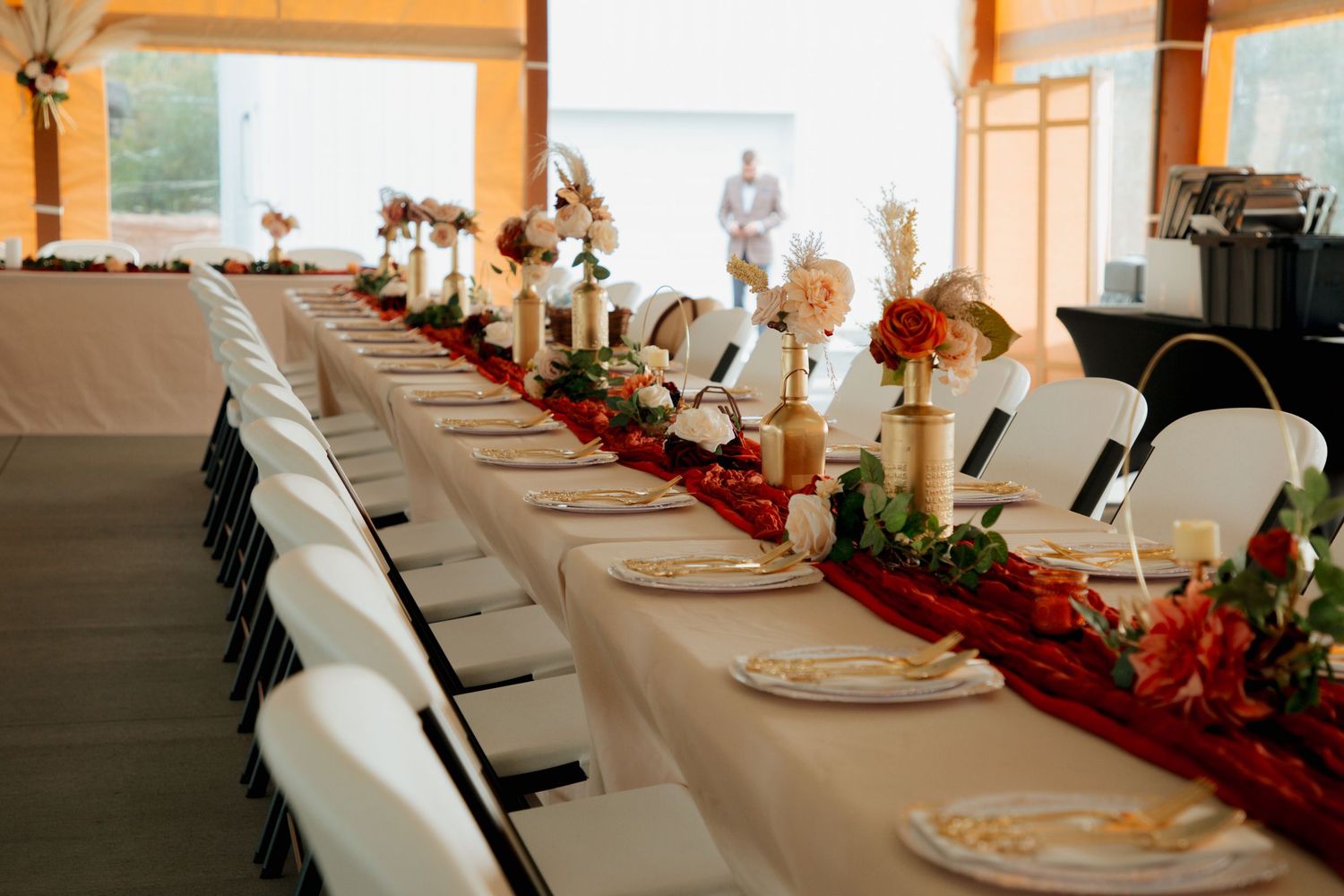 Sweetheart wedding reception table with red velvet runner, gold floral arrangements, and white chairs in open venue.