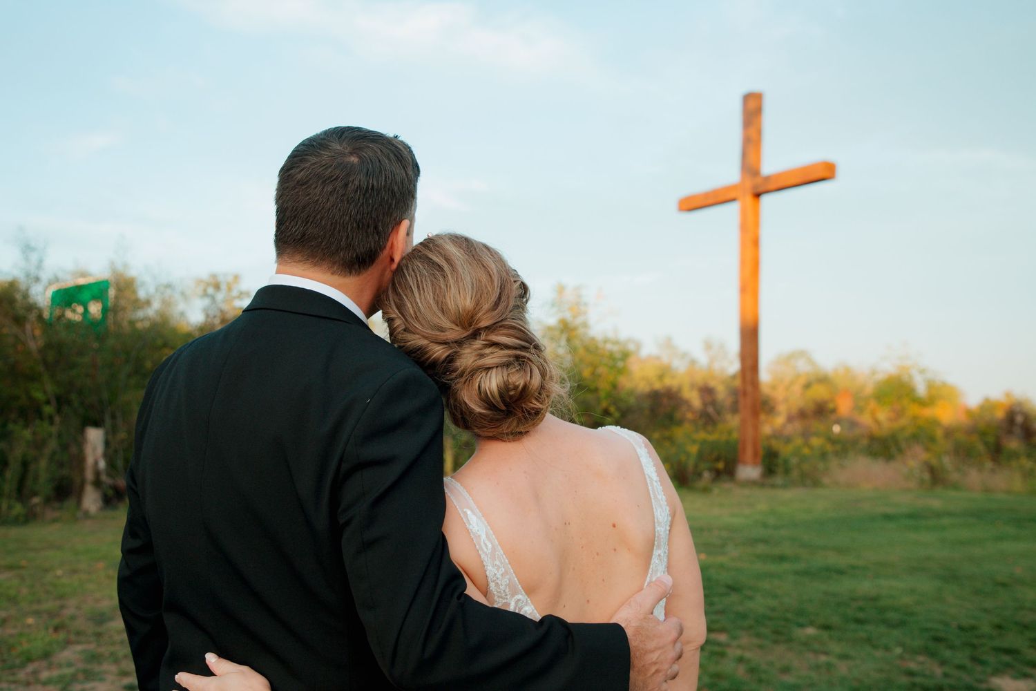Bride resting head on grooms shoulder as they face a large wooden cross at sunset.