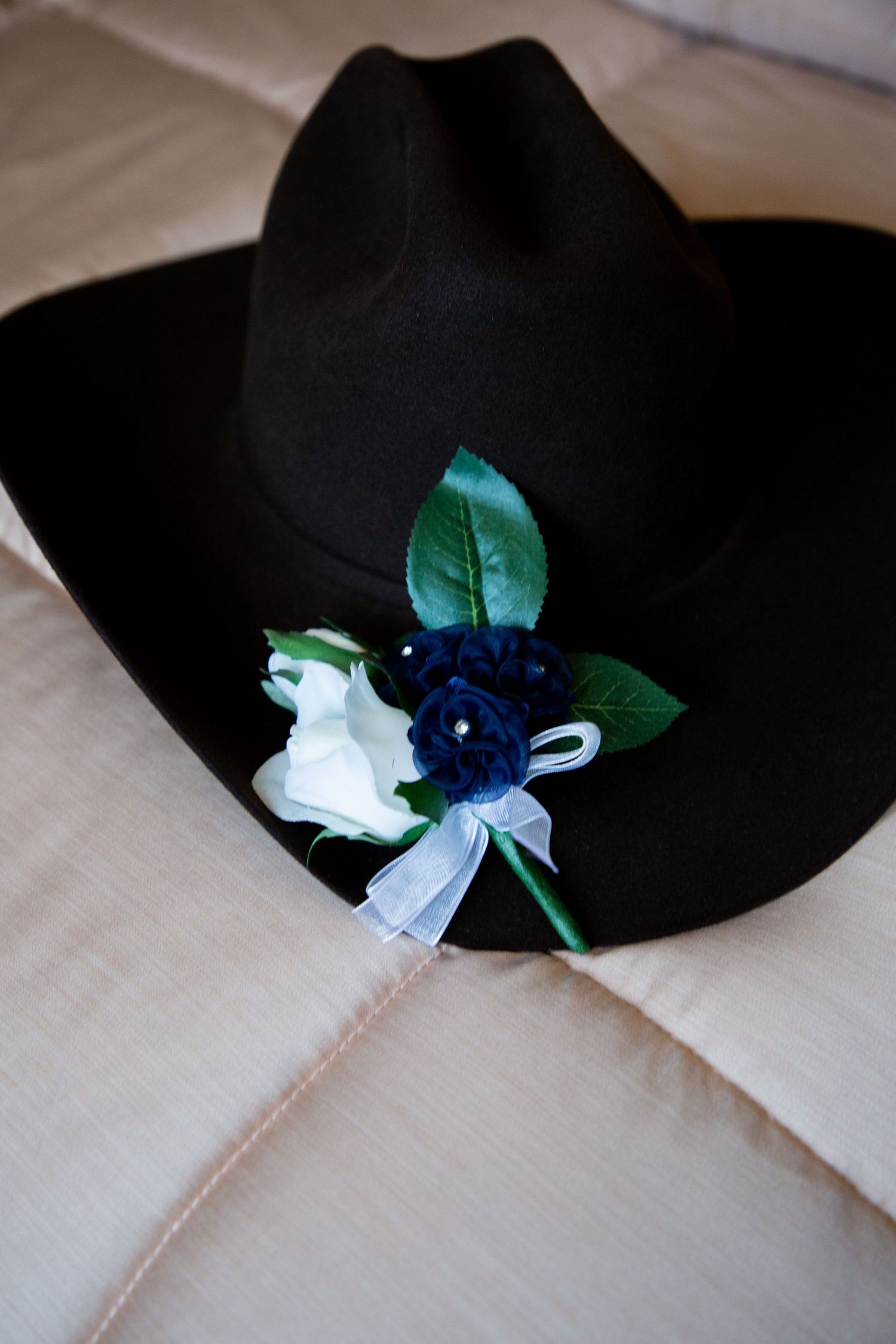 Black cowboy hat adorned with white and navy blue floral boutonniere on beige fabric.