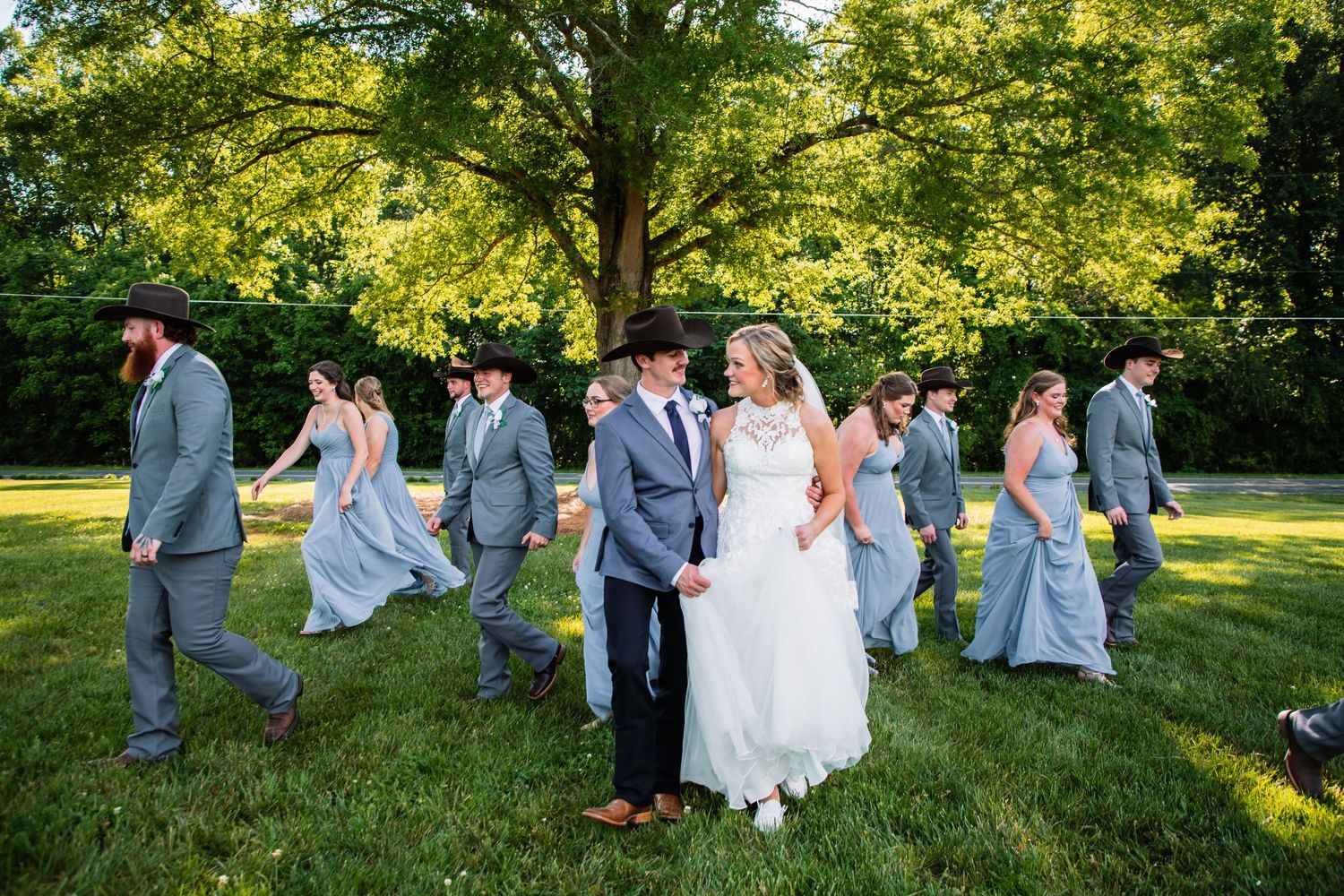 Bride and groom lead wedding party in a fun walk across green lawn in golden afternoon sunlight.