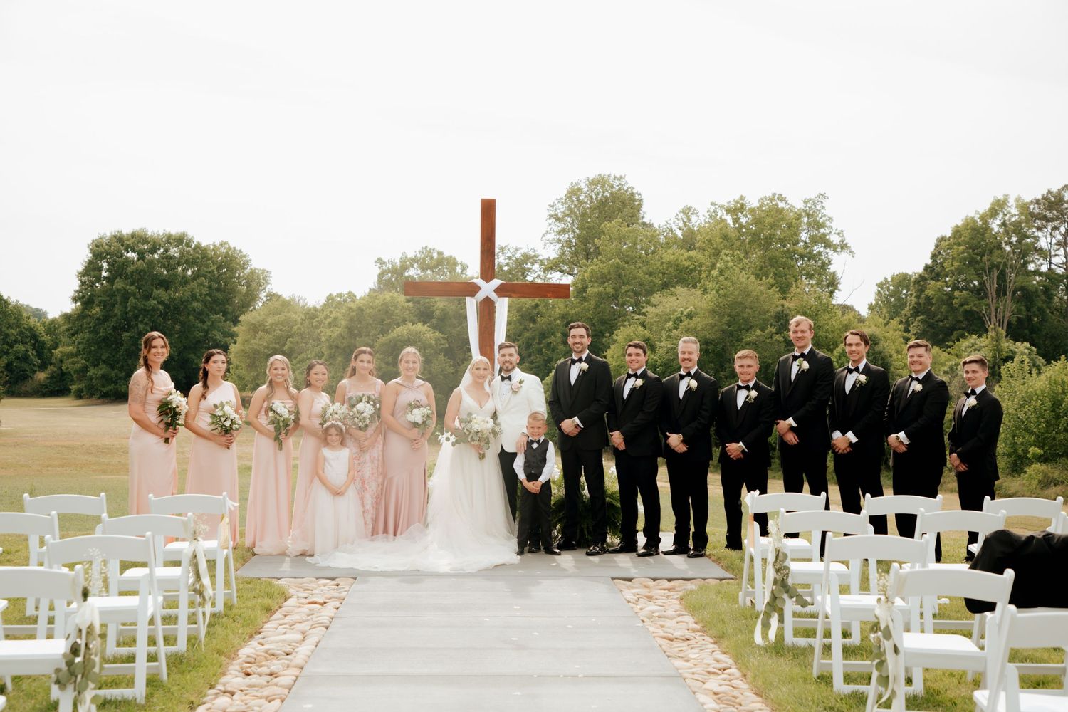 Wedding party posing in front of wooden cross at outdoor ceremony with white chairs.