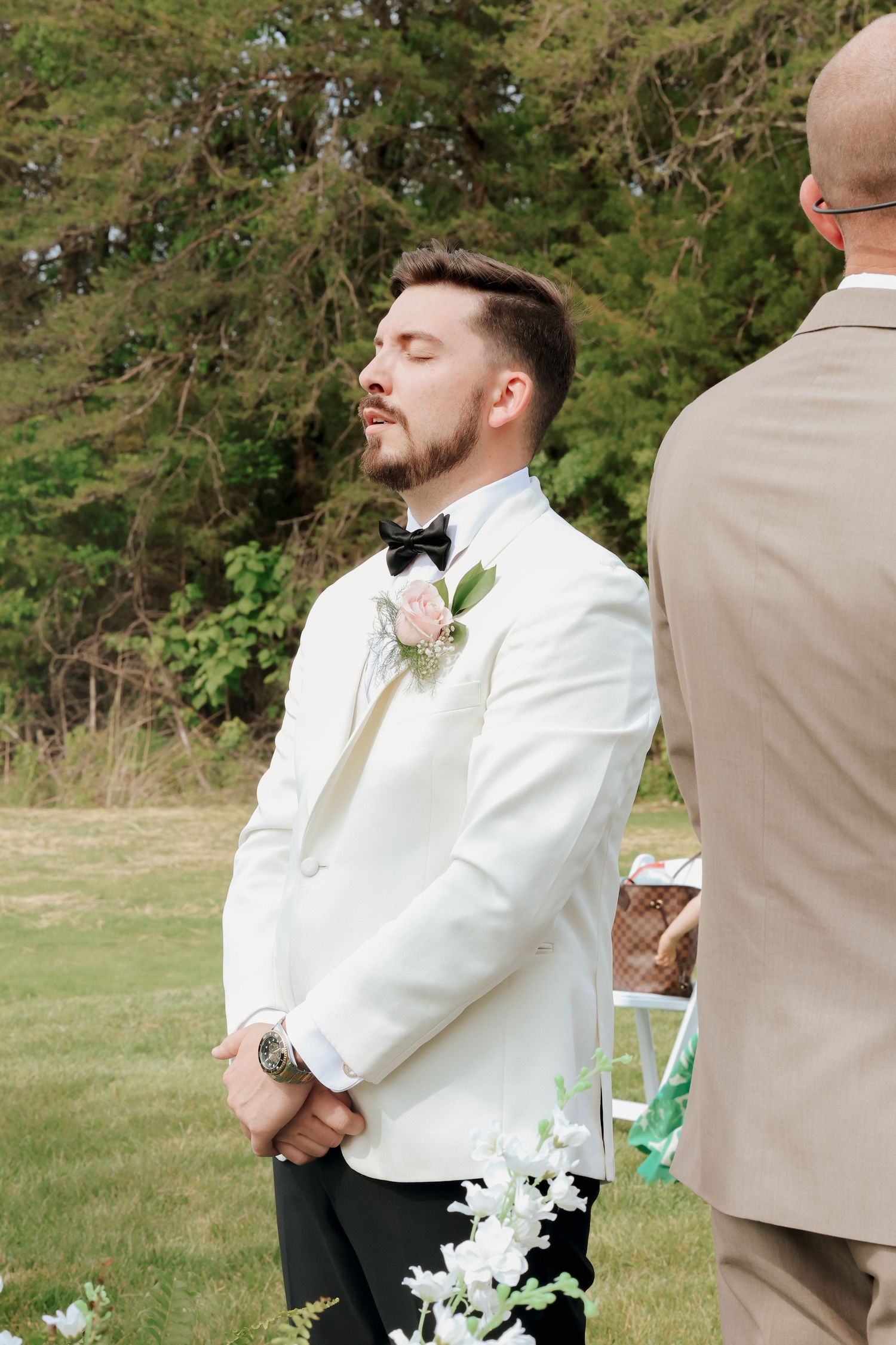 Emotional groom in white tuxedo jacket with eyes closed awaiting bride at outdoor wedding ceremony.