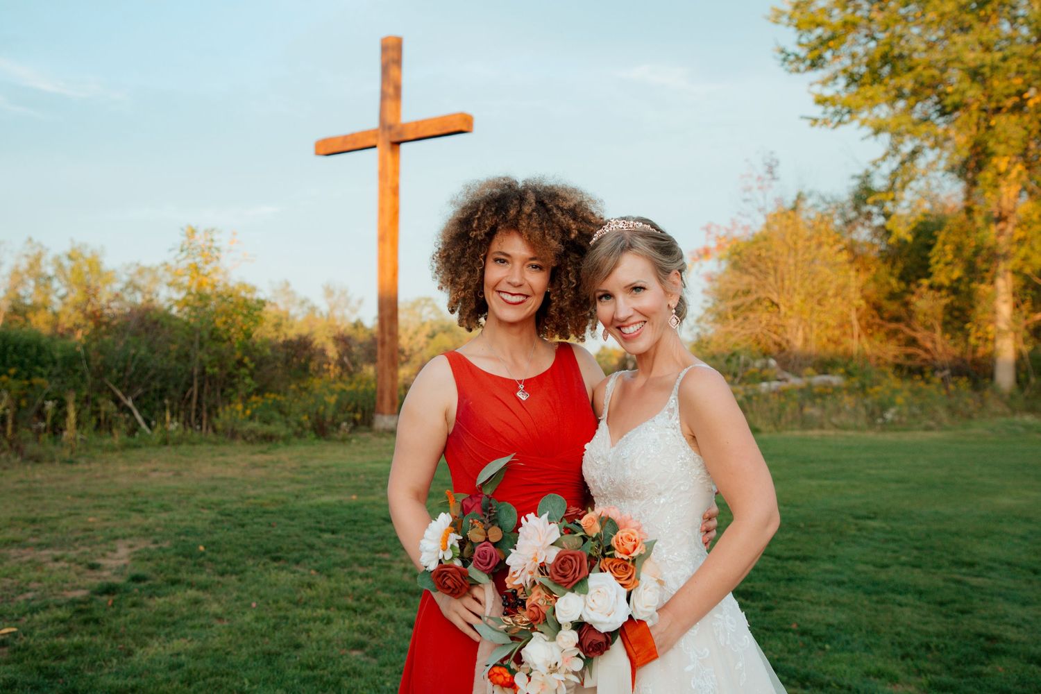 Bride in lace gown posing with a friend in red dress near a wooden cross outdoors.