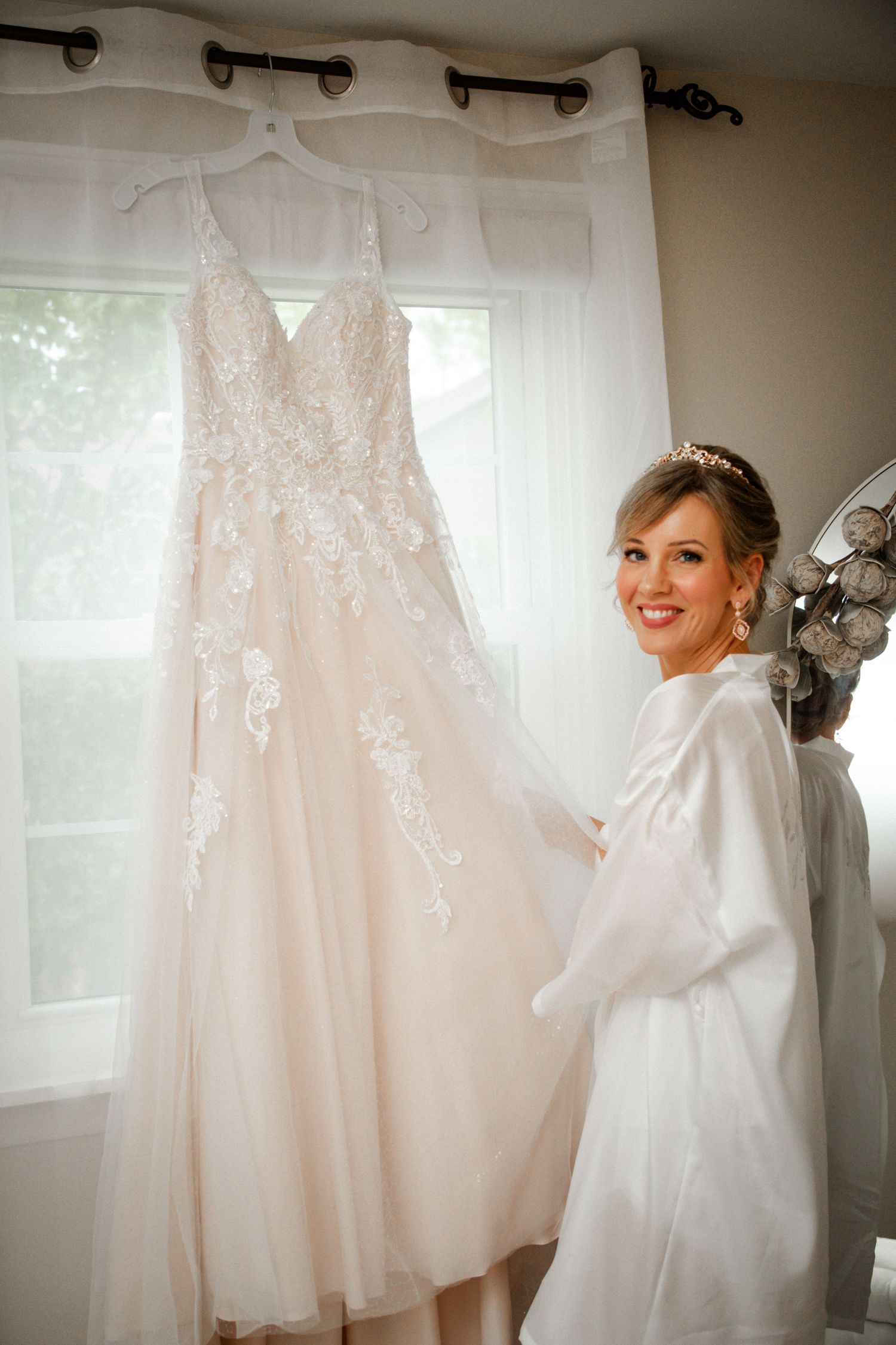 Smiling bride in white robe posing beside her blush lace wedding gown.