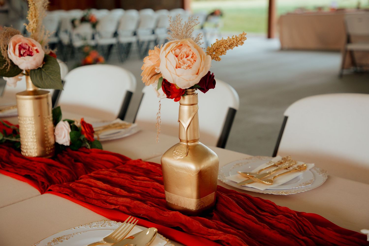 Wedding reception table with gold painted vases holding peach and cream flowers, red velvet runner, and gold place settings.
