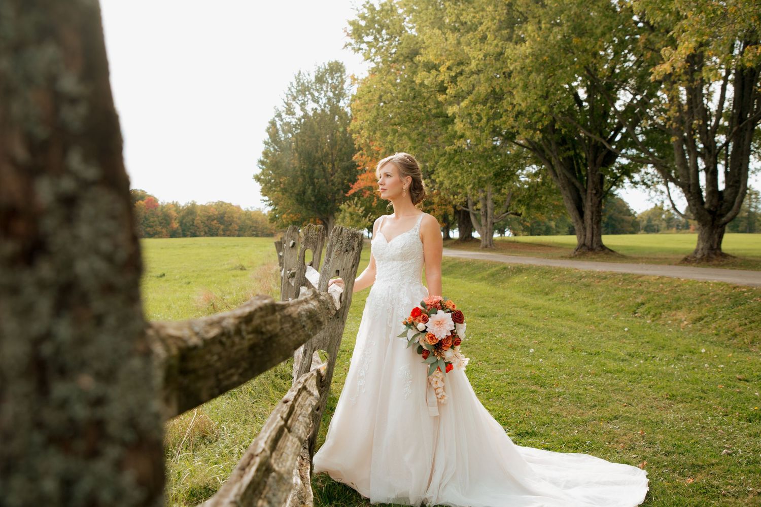 Bride in a flowing white gown holding an autumn bouquet standing by a rustic fence in a sunlit field.