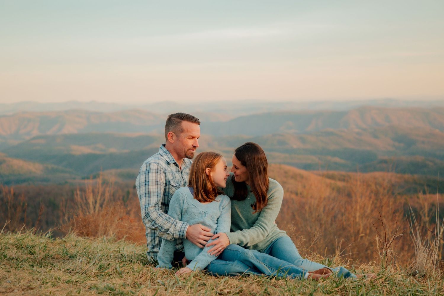 A family of three sits closely together on a grassy mountain overlook, sharing a tender moment at golden hour.