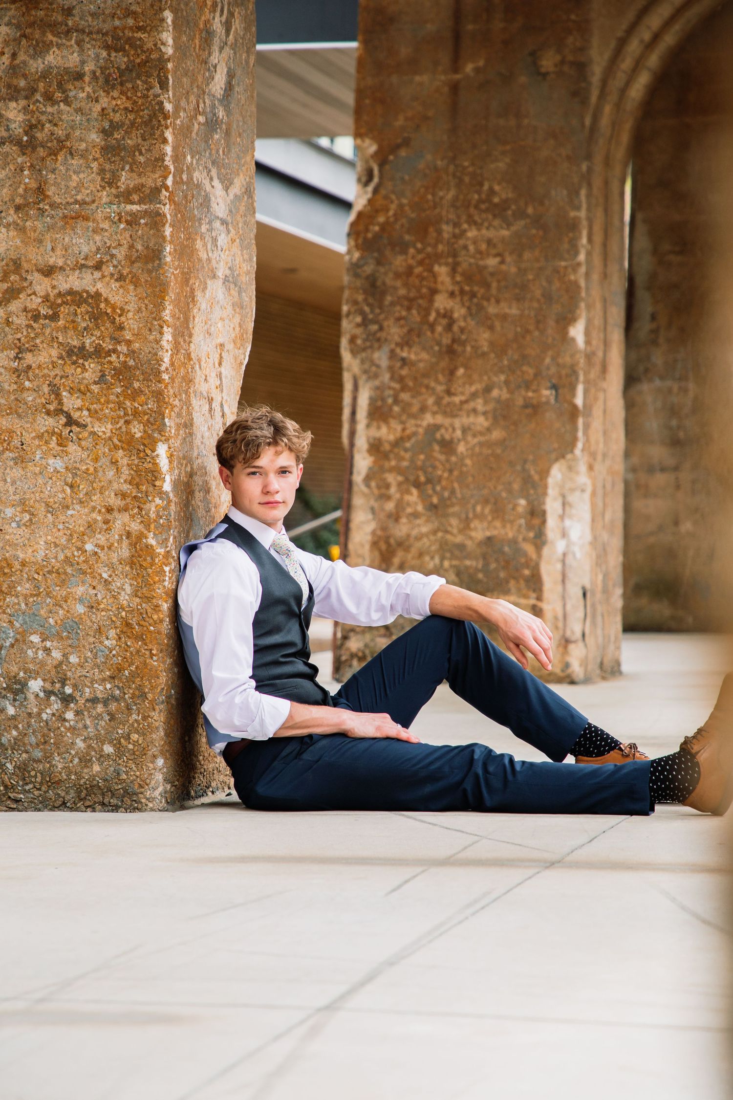 Young man in formal vest and tie sits relaxed against ancient stone pillars on tile floor.