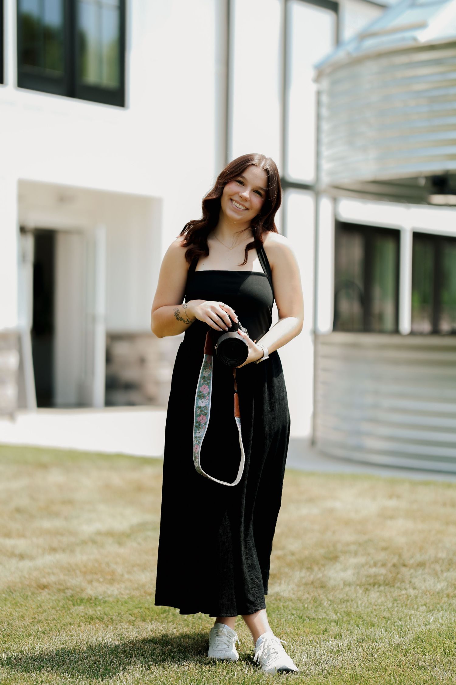 Smiling brunette woman in black sundress holds camera with floral strap while standing on grass.