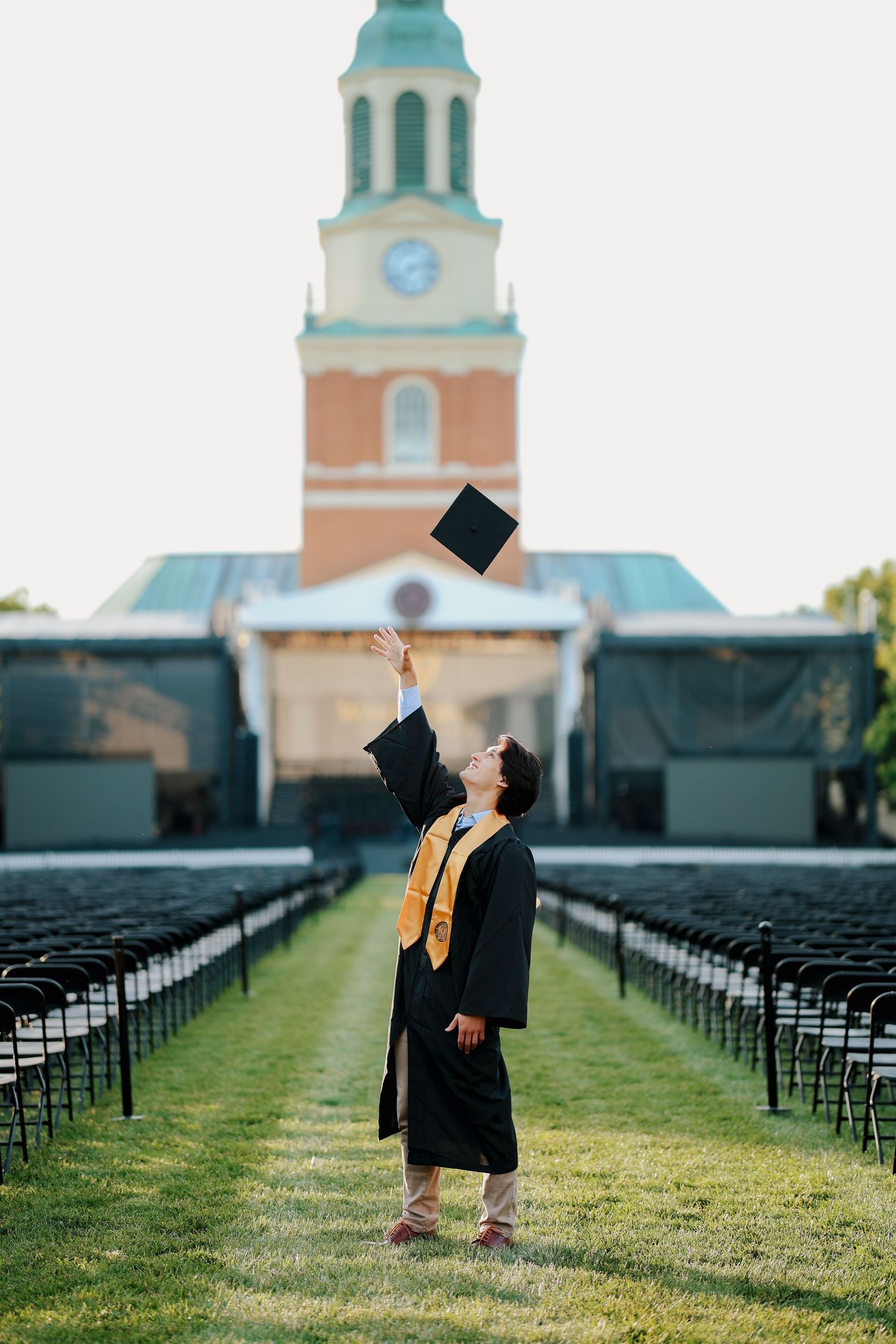 Graduate in black robe tosses cap skyward before a brick chapel with clock tower.