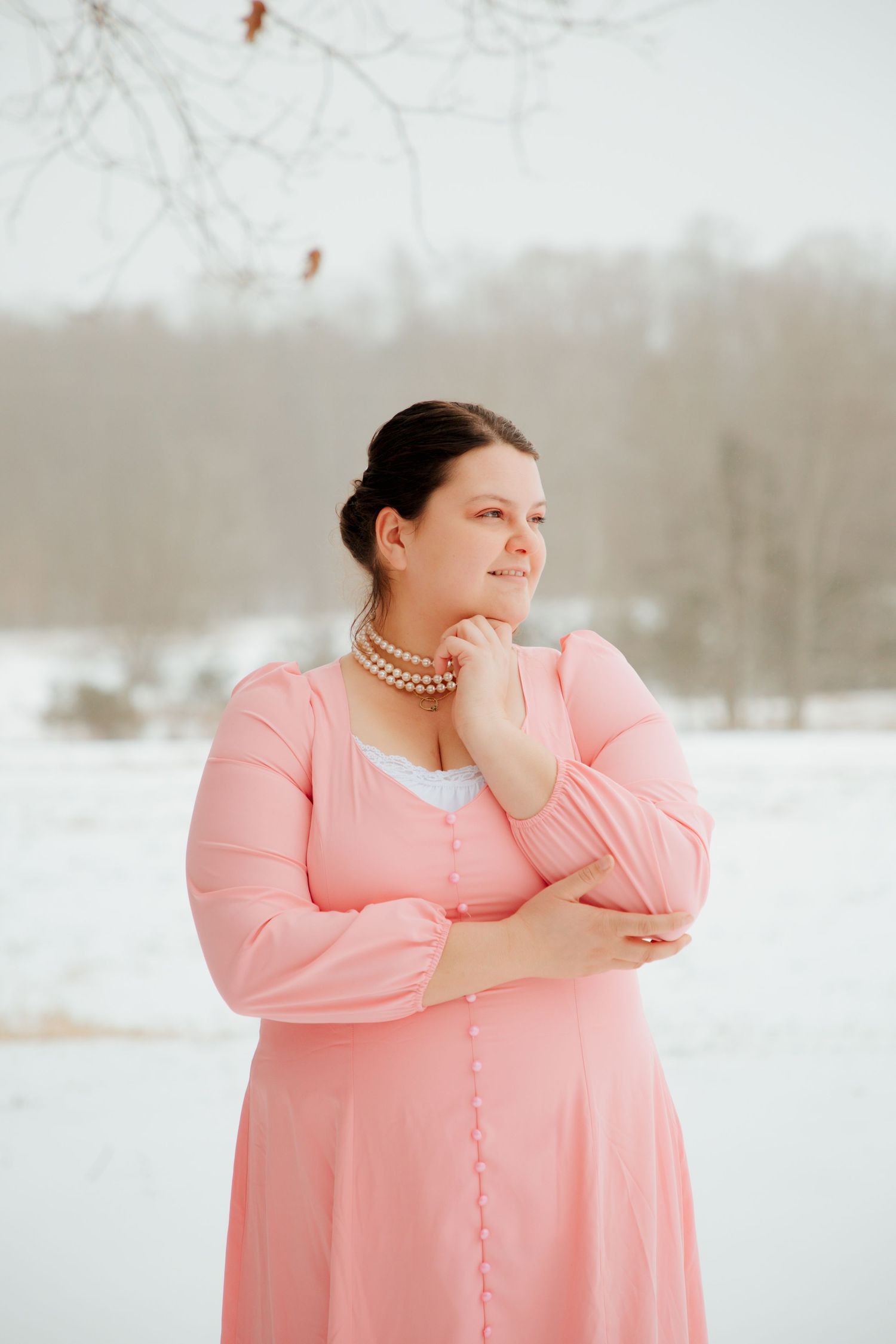 Elegant plus-size woman in pink dress and pearls gazing softly in a winter landscape.