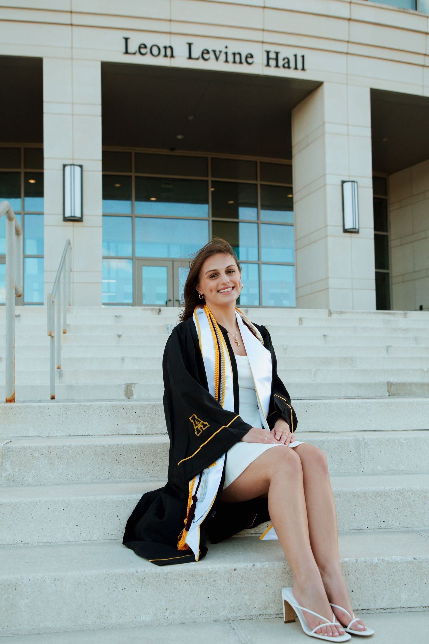 Smiling graduate in black gown sits on steps outside Leon Levine Hall building.